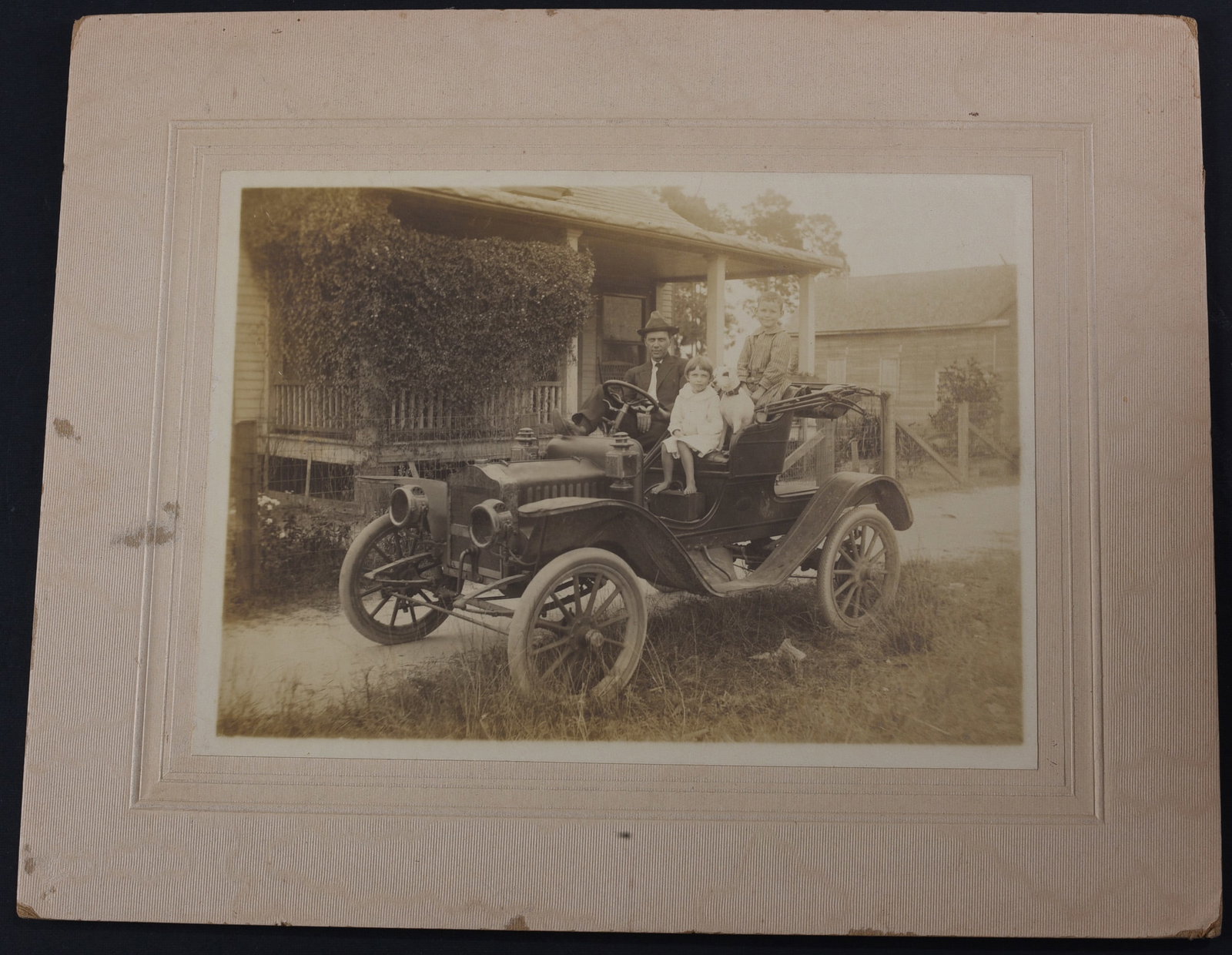 1910 Automobile Photograph: Albumen photograph 7”x5” of a man, his daughter, and their dog in an early automobile. C. 1910. Mounted to period paper mount. Very good condition.
