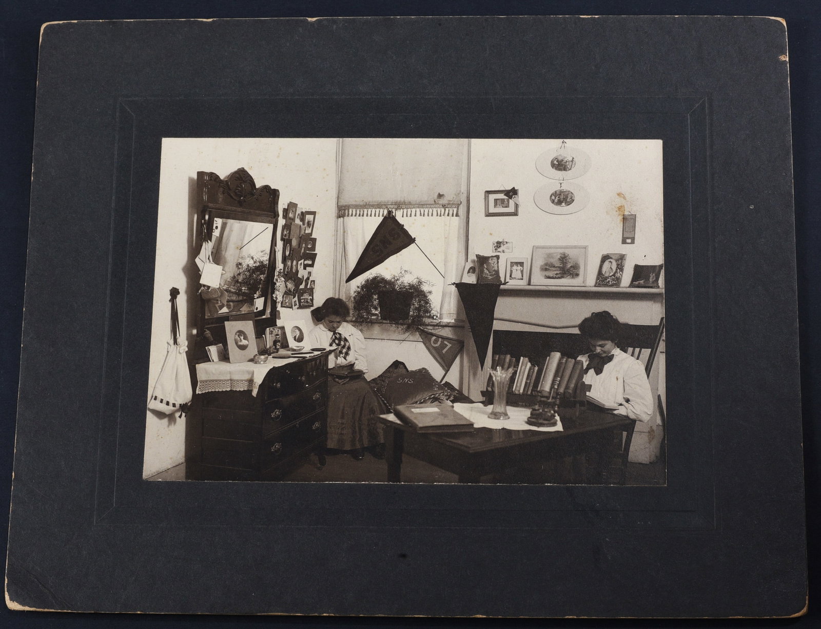 1907 College Dorm Life: Albumen photograph 7”x5” of two women in a college dorm setting. “SNS” pennant and pillow visible, and “07” pennant in view. Mounted to period board. Very good condition.