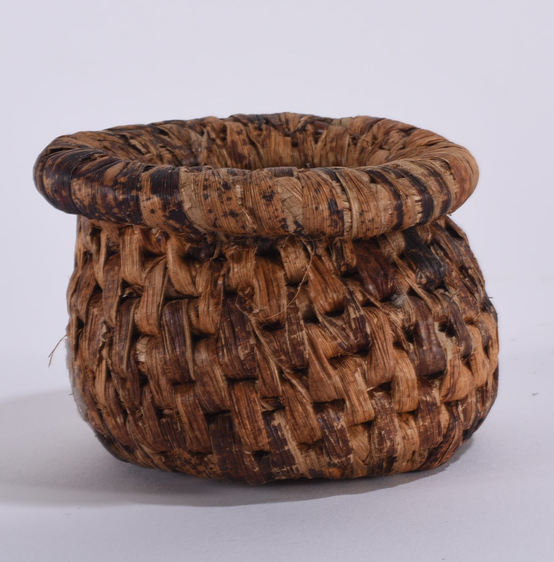 Miniature Basket: Small handwoven miniature basket of natural plant fiber, tightly constructed with a rounded body, thick wrapped rim, and warm brown coloration. Displays a pleasing irregularity and visible
