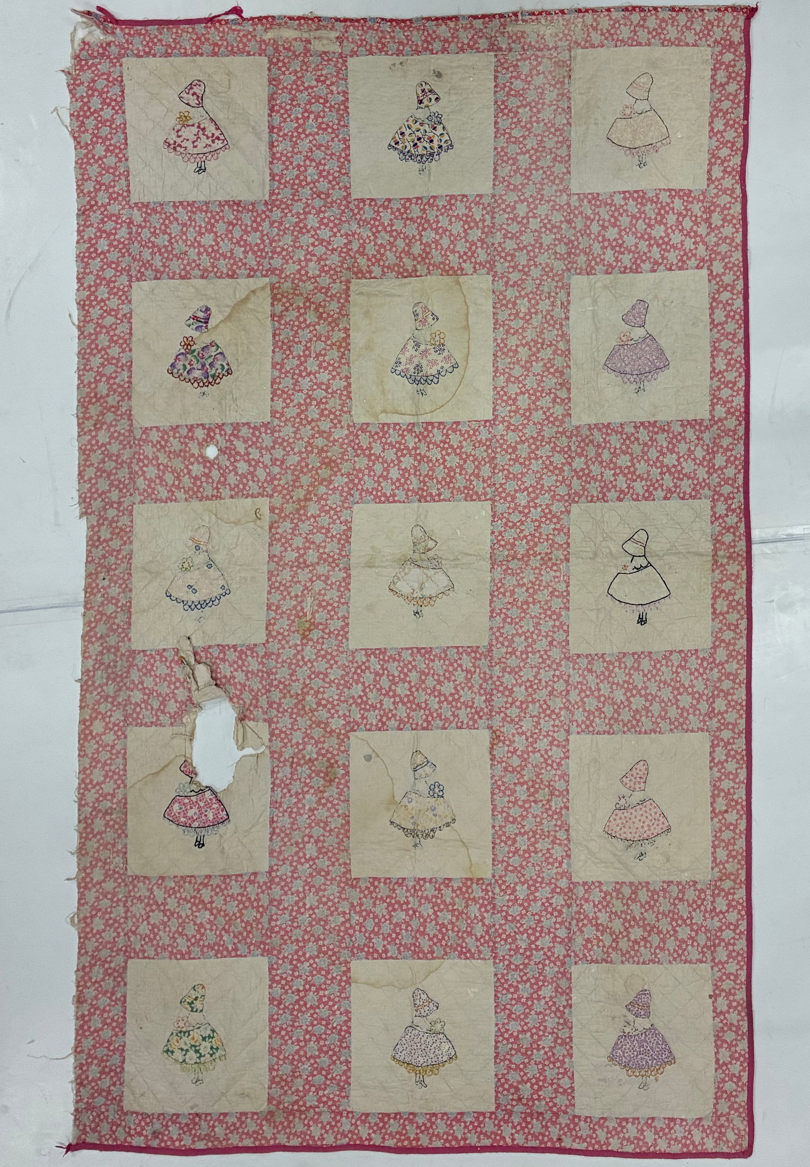 American Sunbonnet Sue Appliqué & Embroidered Quilt, Circa 1930-1940: 82"x50" Condition issues see photos. Charming American Sunbonnet Sue quilt featuring fifteen individual bonneted girl figures worked on cream square grounds and separated by broad pink floral-print sa