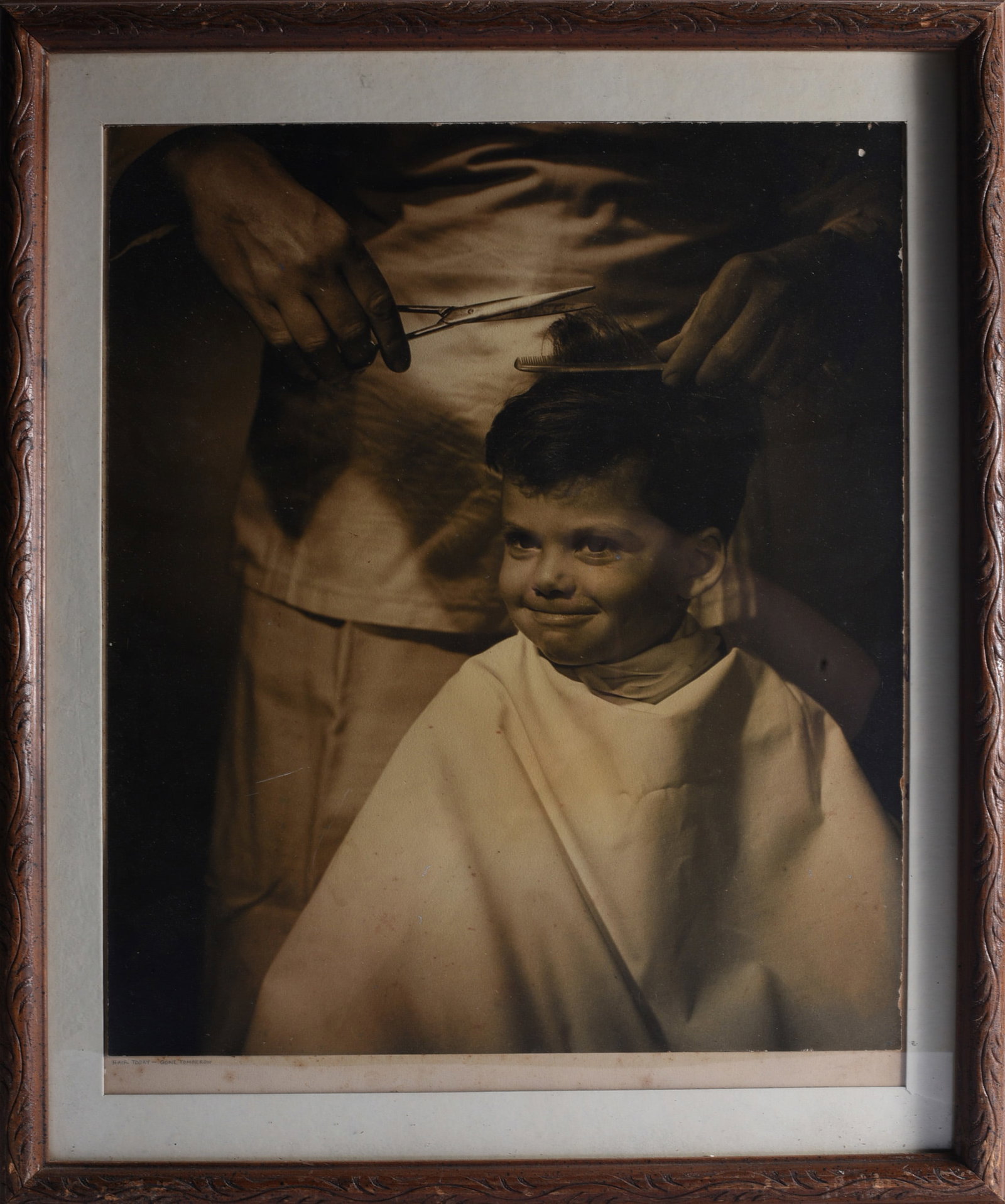 Original Barber Shop Photograph: 18"x22" Original vintage barber shop photograph depicting a young boy seated beneath a barber’s cape while his hair is being cut, with the barber’s hands, comb and scissors visible above. The dram