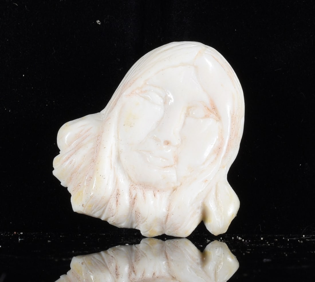 Inuit Carved Bone Female Portrait: 1" Inuit carved bone female portrait depicting a woman with long hair framing the face. Hand carved with softly modeled facial features, incised hair details and a smooth polished cream-white surface.