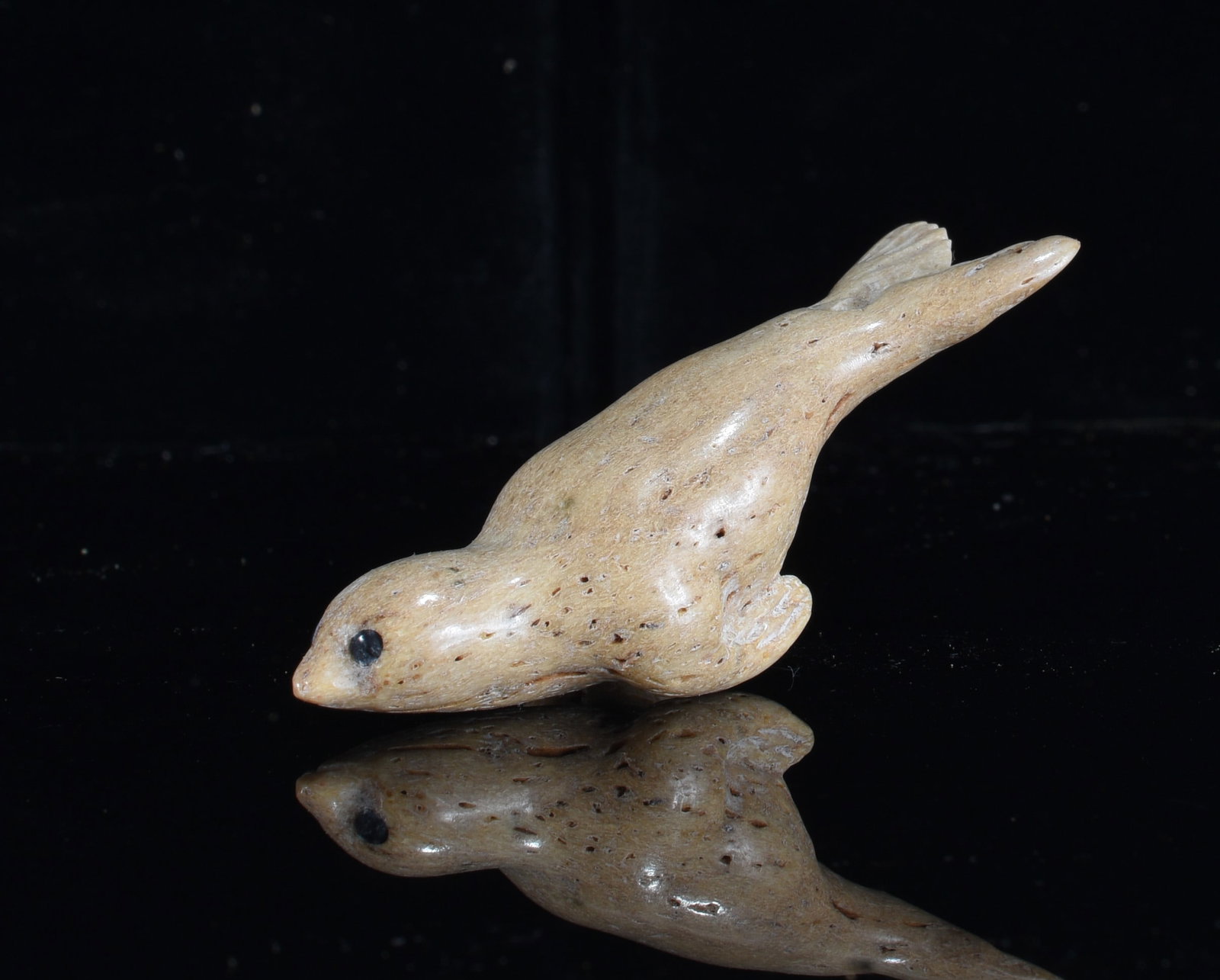 Inuit Carved Bone Seal Figure: 1" Inuit carved bone seal figure depicted in a streamlined swimming pose with dark accented eye, carved flippers and tapered rear body. Warm tan coloration with natural porous surface variations.