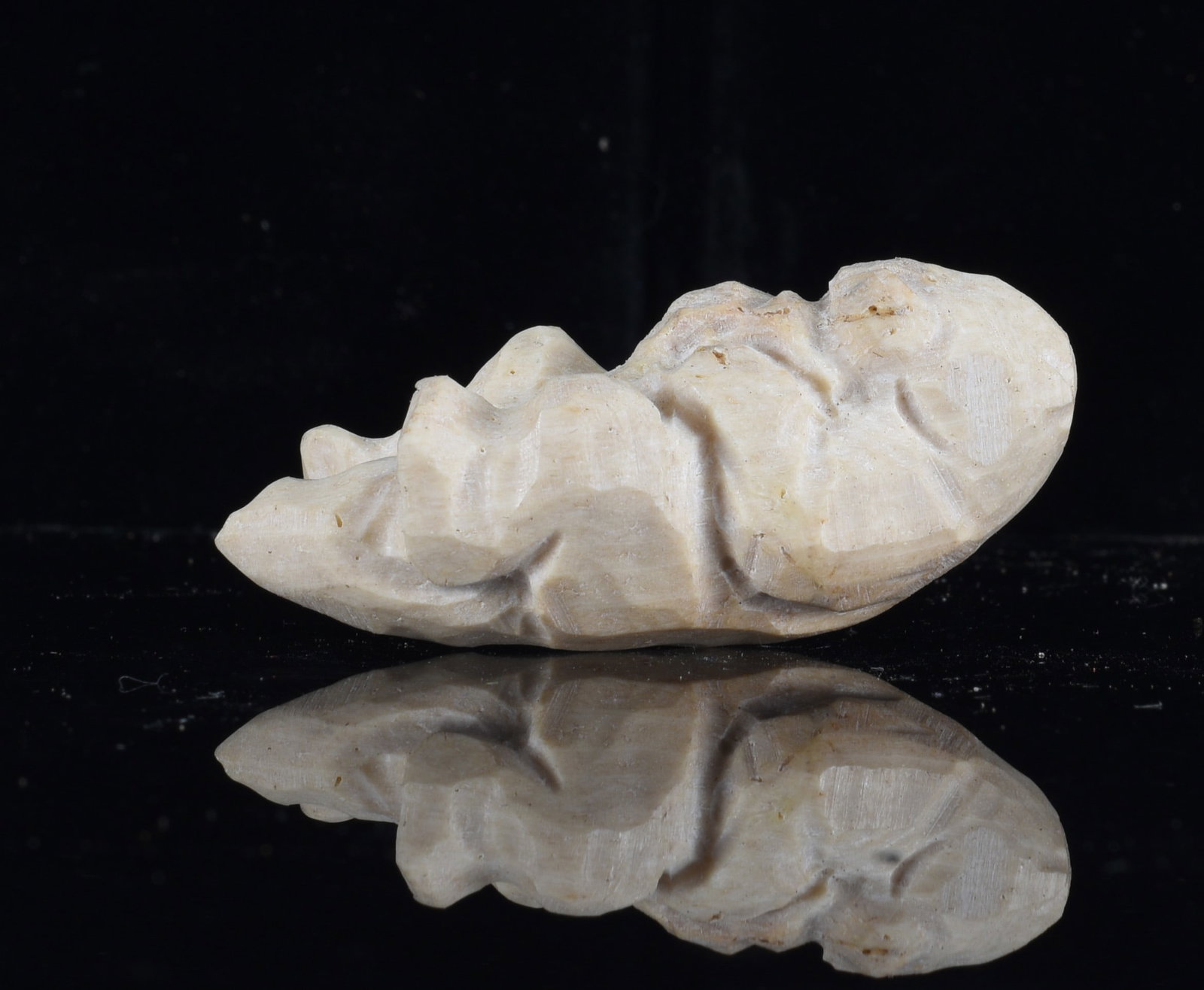 Inuit Carved Bone Reclining Figure: 2"x1" Inuit carved bone reclining human figure shown lying on its back with bent legs and arms positioned across the body. Simplified hand-carved features with a cream-colored surface and natural vari