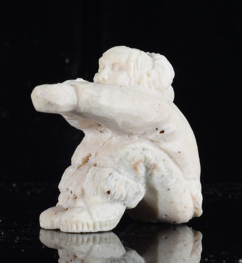 Inuit Carved Bone Seated Hunter Figure: 1.5" Inuit carved bone seated hunter figure wearing traditional cold-weather clothing with an extended arm and bent legs. Cream-colored surface with incised garment details, natural variation and age-