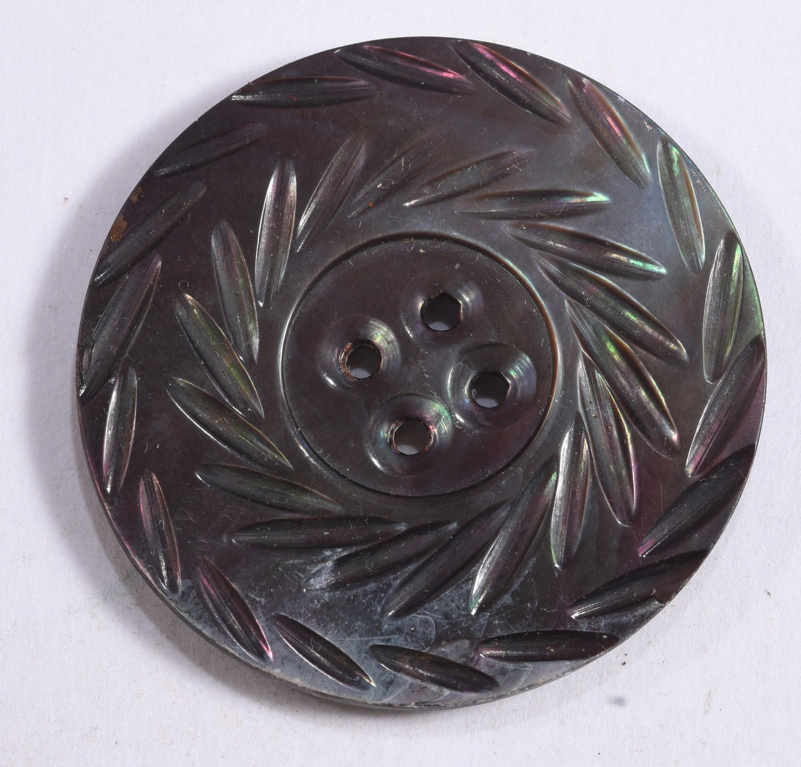 Large Antique Carved Iridescent Composition Button: Large round antique dark composition button with four central sewing holes and deeply molded elongated leaf decoration arranged around the face. The surface displays subtle purple, green and reddish i