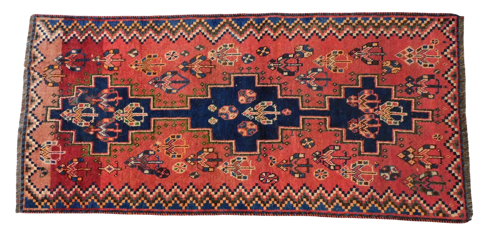 RUG: Qashqai, 2' 6" x 5' 4", wool on wool, red ground, navy, tan, and orange accents, repeating: RUG: Qashqai, 2' 6" x 5' 4", wool on wool, red ground, navy, tan, and orange accents, repeating bow/arrow and shield motifs, wear consistent with age and use including areas of wear to ground.