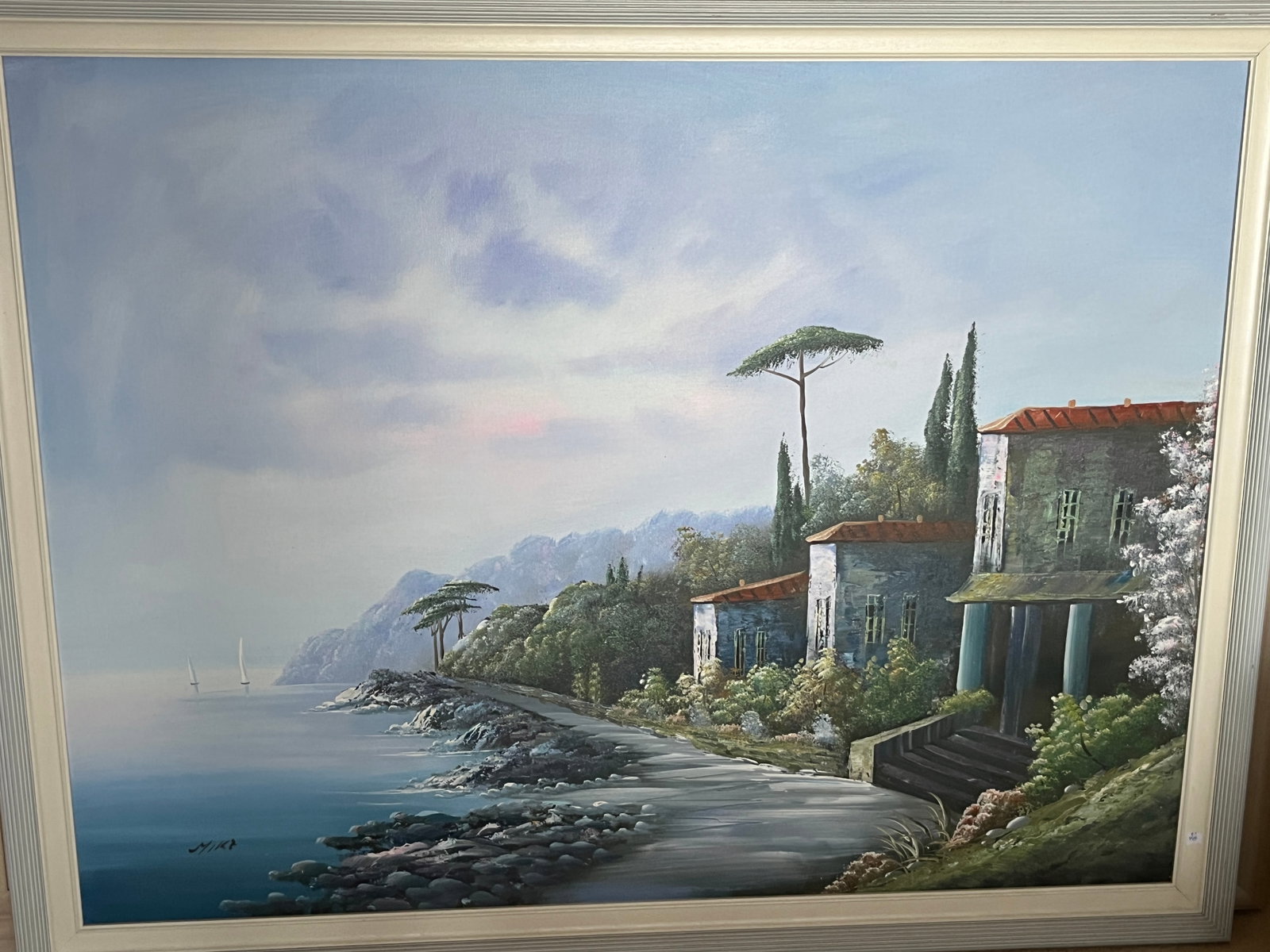 Mika (Contemporary) – Mediterranean Coastal Villas, 36x48 Oil on Canvas: A serene coastal landscape depicting a row of Mediterranean-style villas with terracotta roofs overlooking a tranquil sea. Two sailboats drift in the distance beneath a soft, clouded sky, while cypres