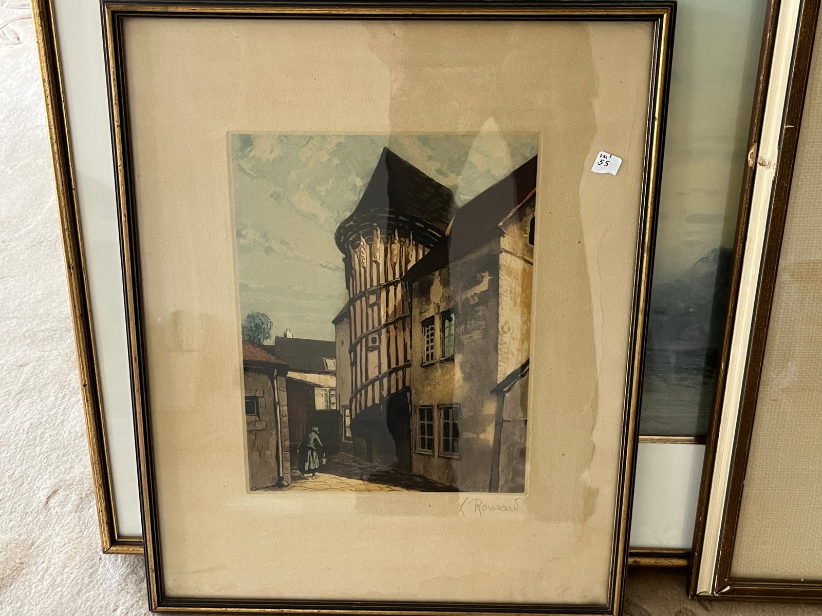 Signed "K. Rousard" European Street Scene Print, Framed 19.5" x 15": Decorative signed print depicting a charming European village street with a prominent half-timbered medieval building and a lone figure walking along a cobblestone lane. Executed in muted earth tones 