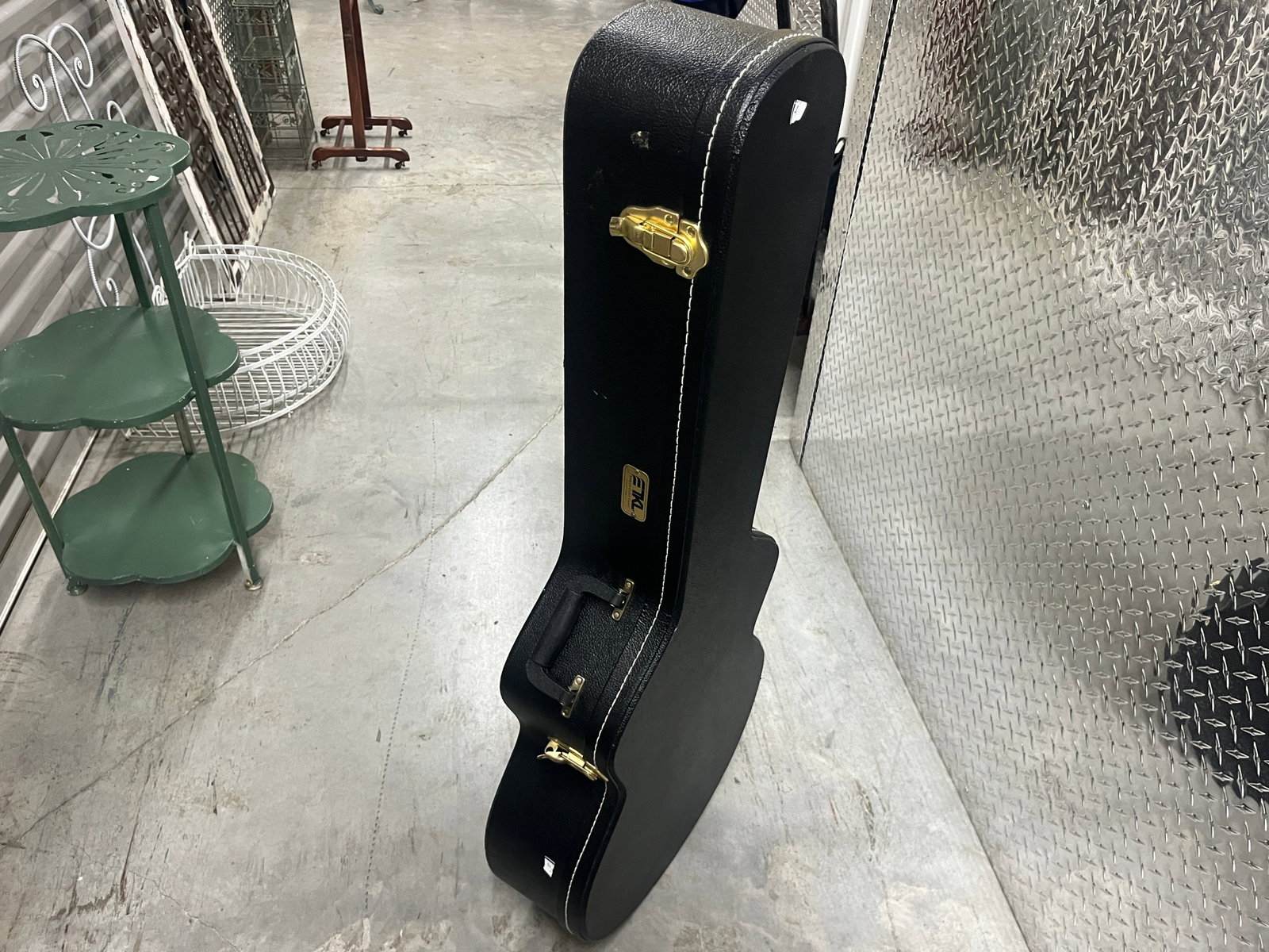 EMG Black Hard-Shell Guitar Case w/ Gray Plush Interior & Brass-Tone Hardware: EMG hard-shell guitar case featuring a black textured exterior with contrasting white stitching, gold/brass-tone latches and hardware, carrying handle and EMG logo badge. Interior is fully fitted with