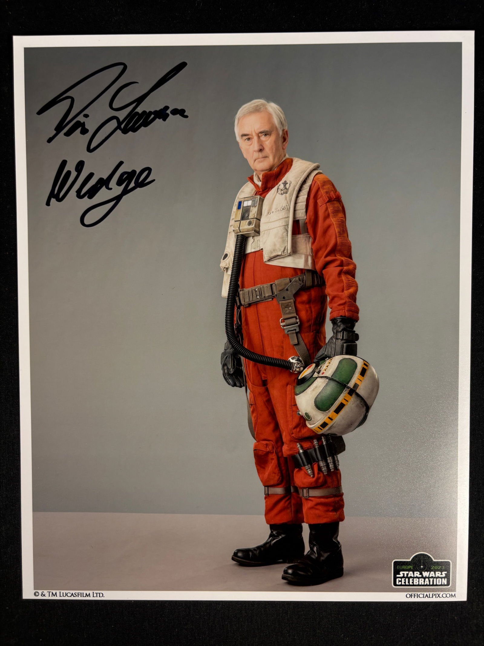 Denis Lawson Signed Star Wars Celebration Europe 2023 Wedge Antilles Photograph: A color photographic print featuring actor Denis Lawson as his character Wedge Antilles from the Star Wars franchise. The studio-lit, full-length portrait depicts Lawson in an orange flight suit and p