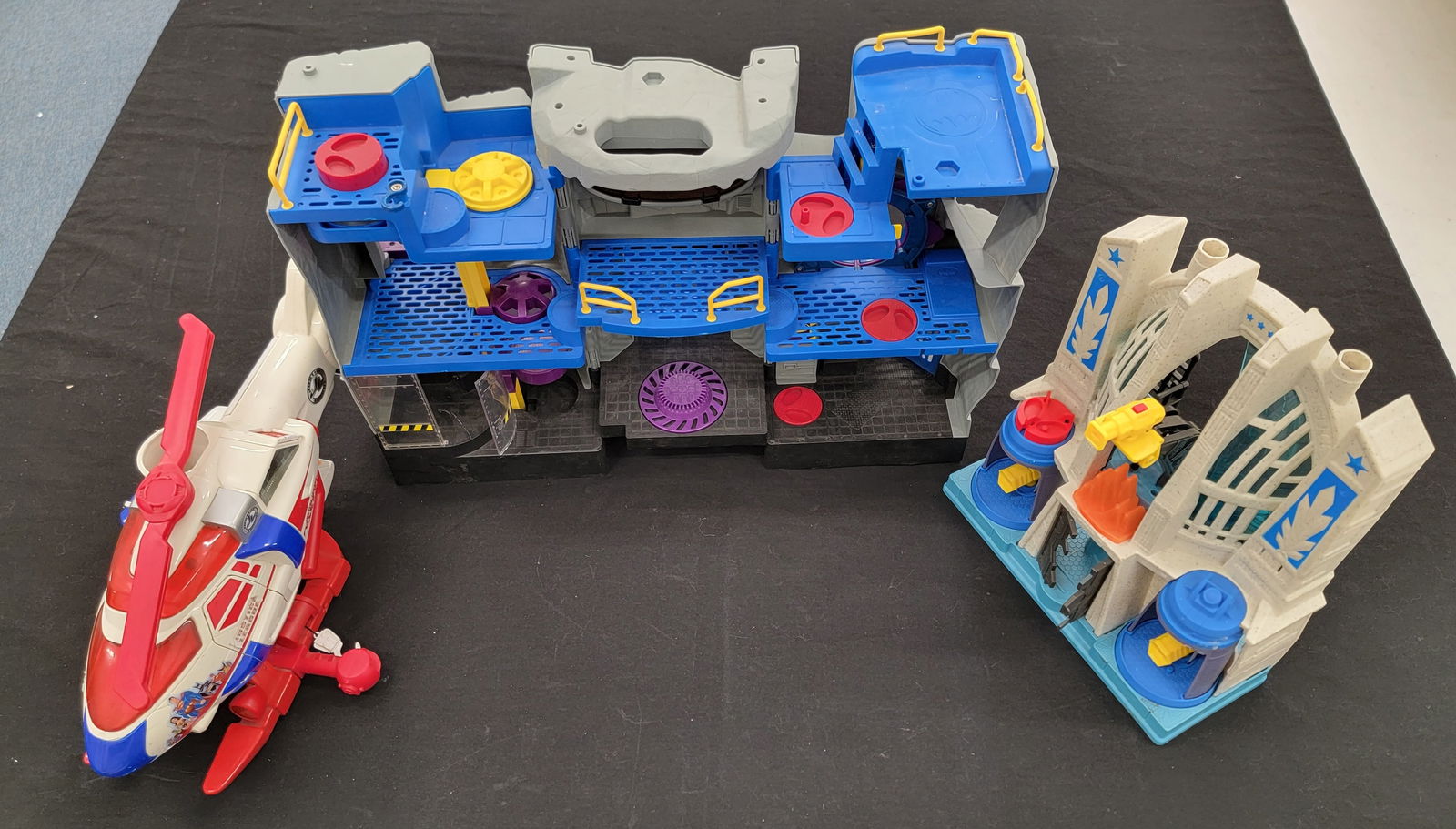 A Trio of Fisher-Price Imaginext DC Super Friends Playsets and a Matchbox Helicopter: A trio of molded plastic playsets and a toy vehicle featuring characters from the DC Comics universe. The group includes a Fisher-Price Imaginext Batman Batcave playset, featuring a gray rocky exterio
