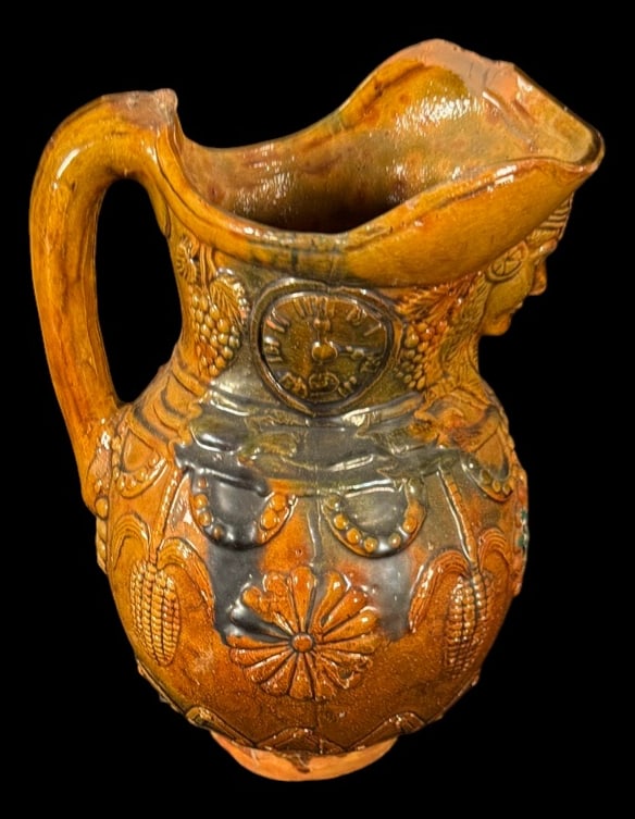 Figural Majolica Style Pottery Pitcher: Decorative glazed pottery pitcher with a classical female face, floral medallions, grape clusters, and embossed decorative motifs in rich brown and green glazes. A colorful carved bird accompanies the