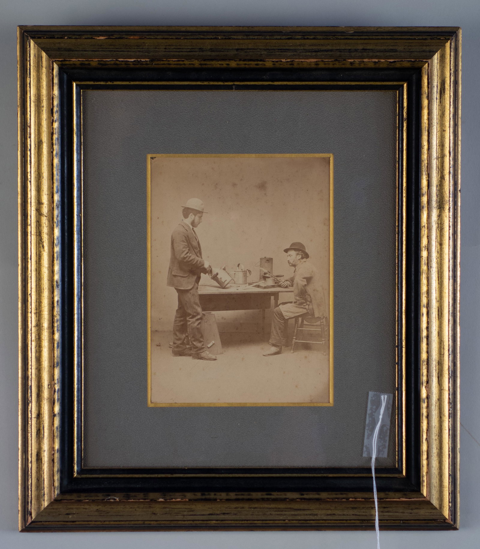 Original Occupational Photograph of Tinsmiths: Original occupational Photograph of two tinsmiths at a table reviewing their wares. One smith is holding a watering can. 5 1/2" x 7 1/2" image size. 13 3/4" x 15 3/4" overall. Framed and matted