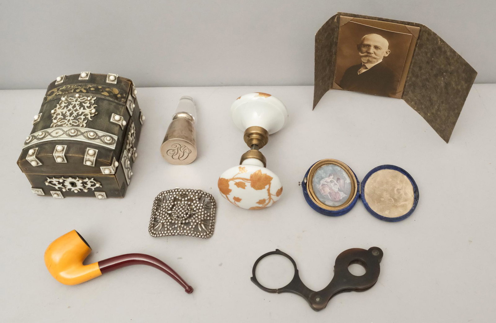 Contents of a Gentleman's Nightstand Drawer: Comprising a folding horn mounted magnifying glass, a pipe, a cased image, a sterling silver decanter top, a porcelain set of doorknobs, a photograph, a steel cut buckle, a horn, ebony, and bone watch
