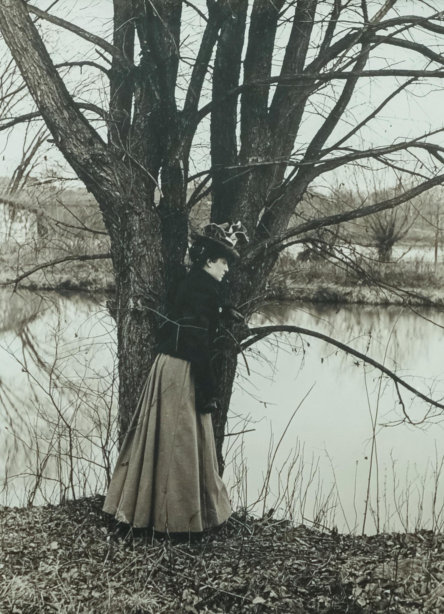 Large Framed Photo of a Victorian Woman: American, late 19th century black and white photograph of a woman bundled against the cold in full skirt, leather gloves, and hat, leaning against a tree and looking absently at the water, housed in a