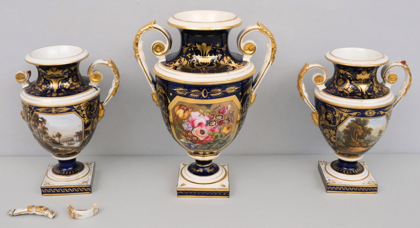Pair of Old Derby Porcelain Landscape Vases: Cobalt field garniture vases with mask handles, scene entitled "On the Dee..." (handles detached and repaired). Together with a larger cobalt vase with floral decoration (cracked handle). 9" and 11 1/