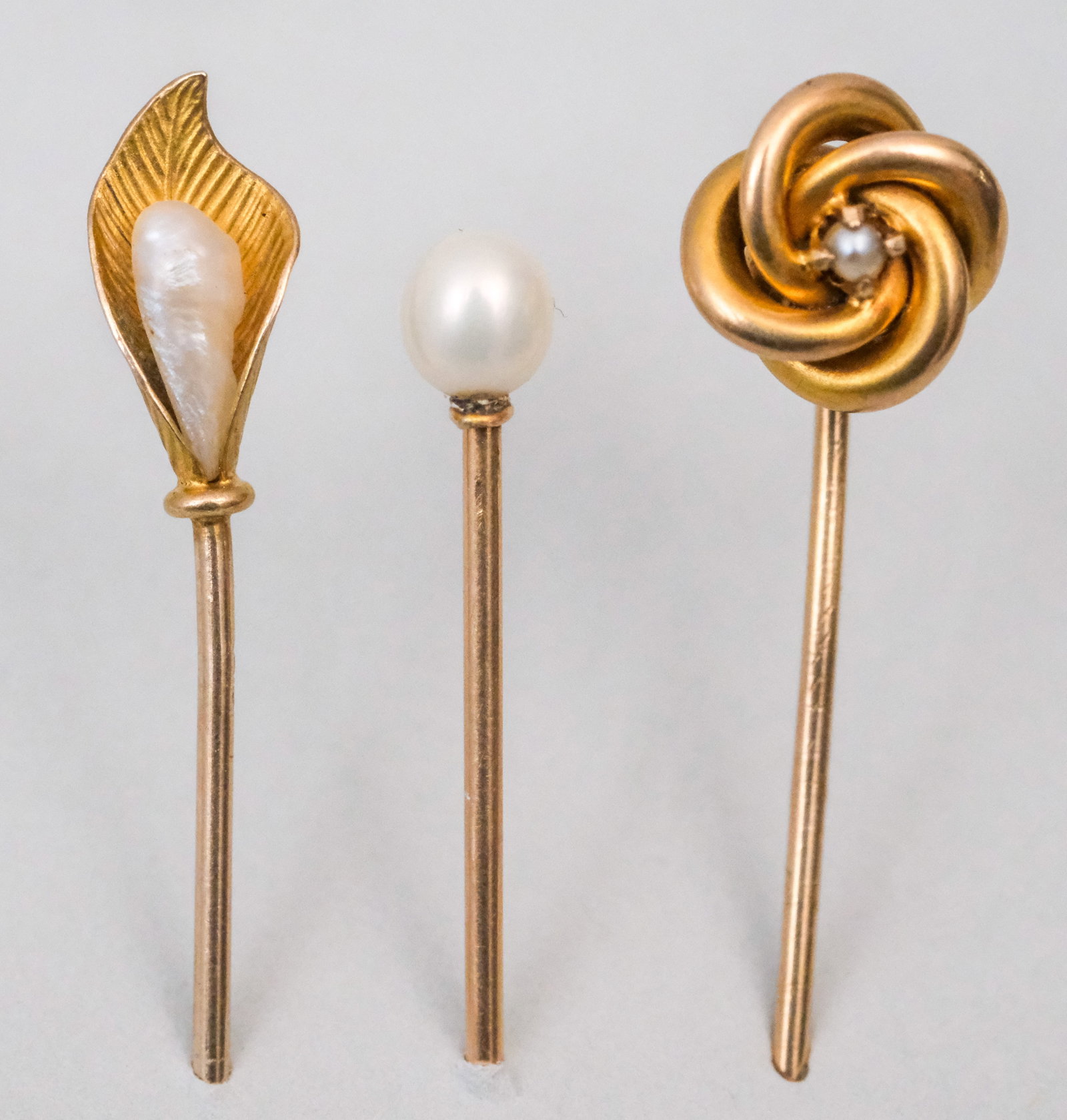 Group of 10K & 14K Gold Stickpins: Victorian Era. Comprising a 10K infinity knot with pearl, a 10K baroque pearl calla lily, and a 14K with pearl head (pearl with hairline). Total weight 10K 1.03 dwt, 14K 0.3. dwt