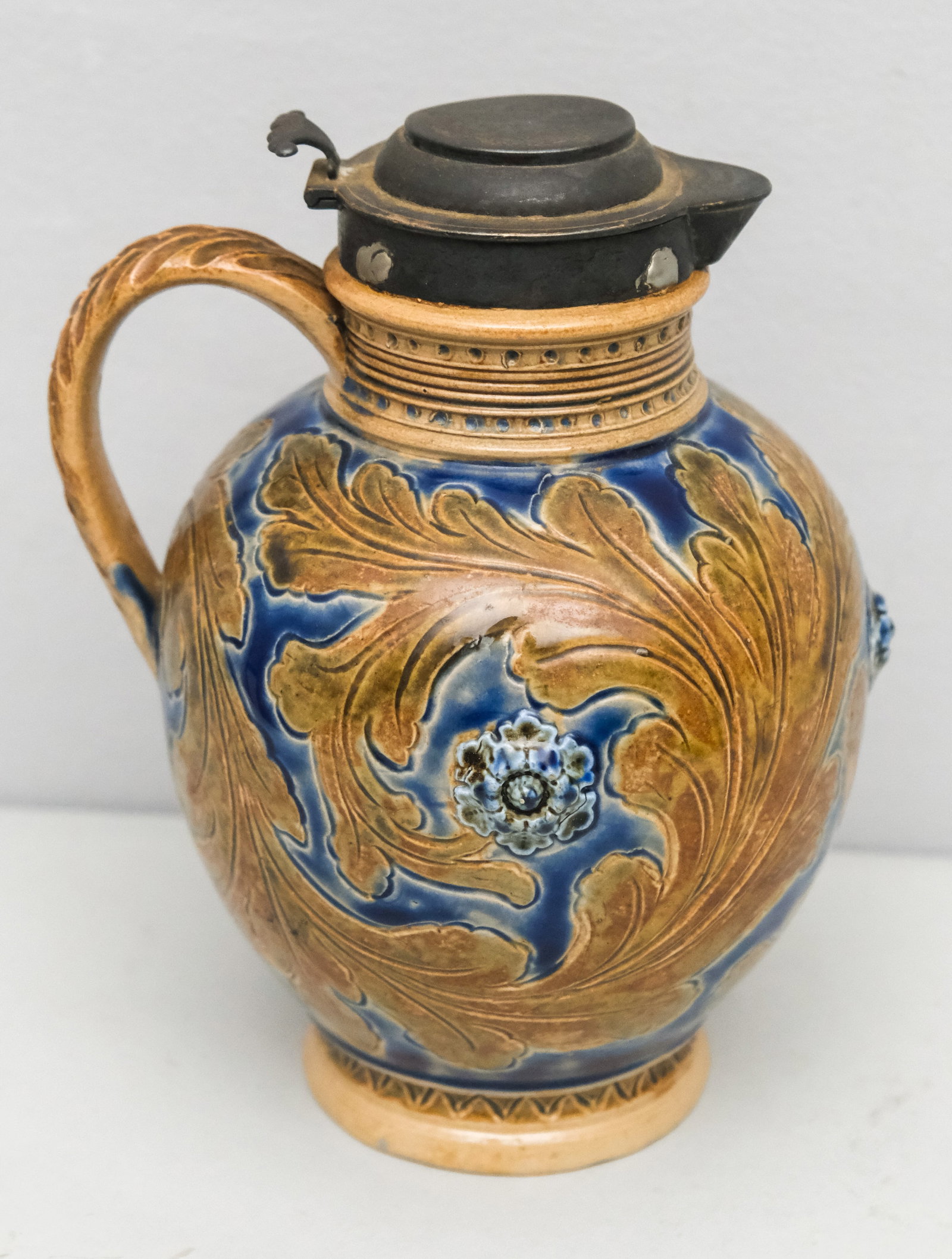 Doulton Lambeth Lidded Pottery Jug: Marked for Arthur Barlow. Large flowing oak leaves over cobalt with rosettes in high relief. Sterling silver collar and lid. 7 1/2"