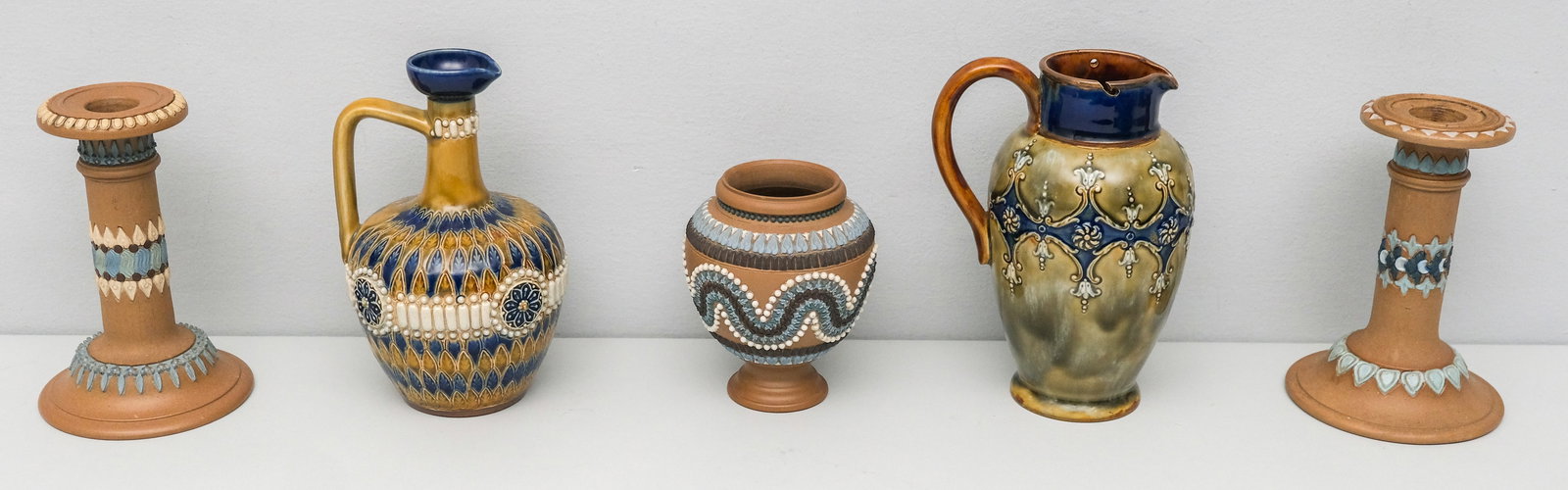 Group of Doulton Lambeth Art Pottery: Comprising a (formerly) lidded pitcher, a cruet (rim chip), a vase (?) (apparently missing lid) and a near pair of candlesticks. Marked as shown. 7" to 4 1/2".