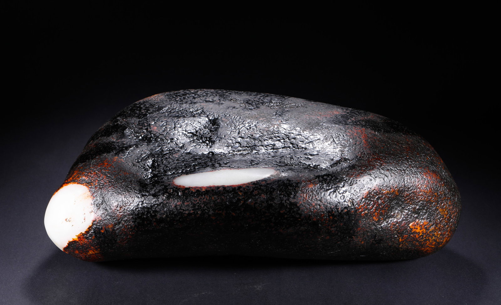 HETIAN JADE BLACK OIL SKIN SINGLE SEED: H:16cm(6.30in) W:38cm(14.96in) Weight:20940g(46.10lb)Top-tier fully wrapped aged black oil skin, with rich deep moist black hue covering the whole stone. The thick original rough patina is fully