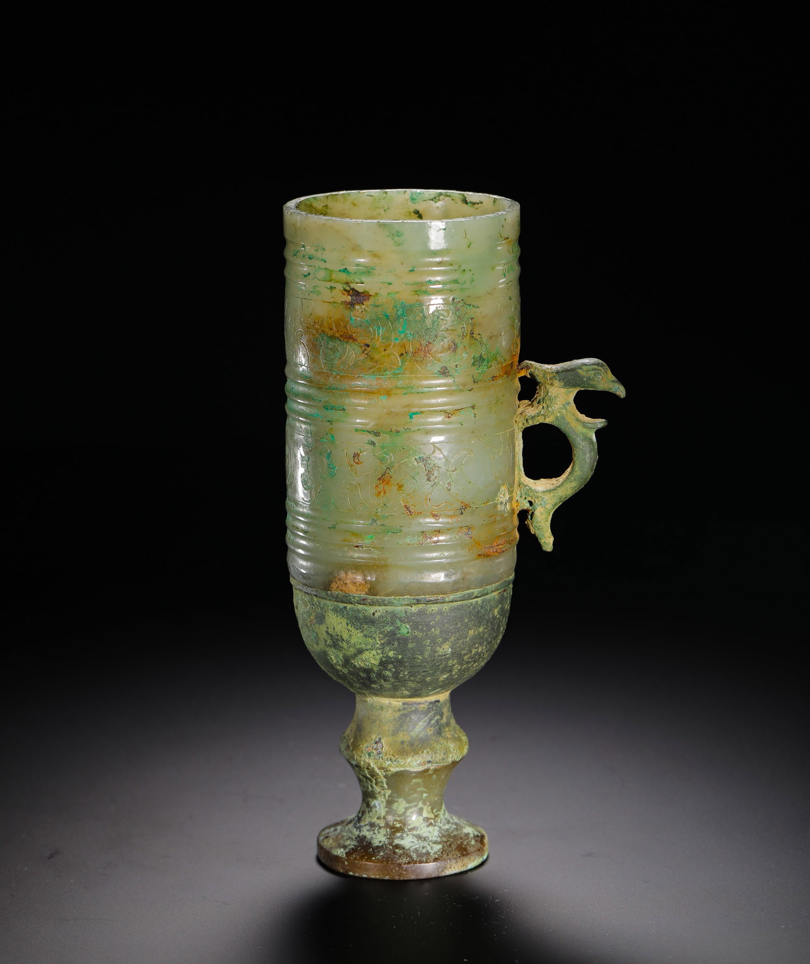 BRONZE BASE JADE PHOENIX EAR CUP OF THE HAN DYNASTY: H:18.3cm(7.20in)W:9cm(3.54in)Weight:580g(1.28lb)This is a Han Dynasty celadon jade cup with a phoenix handle, decorated with raised bands and incised patterns, paired with a bronze high-foot base. Wit
