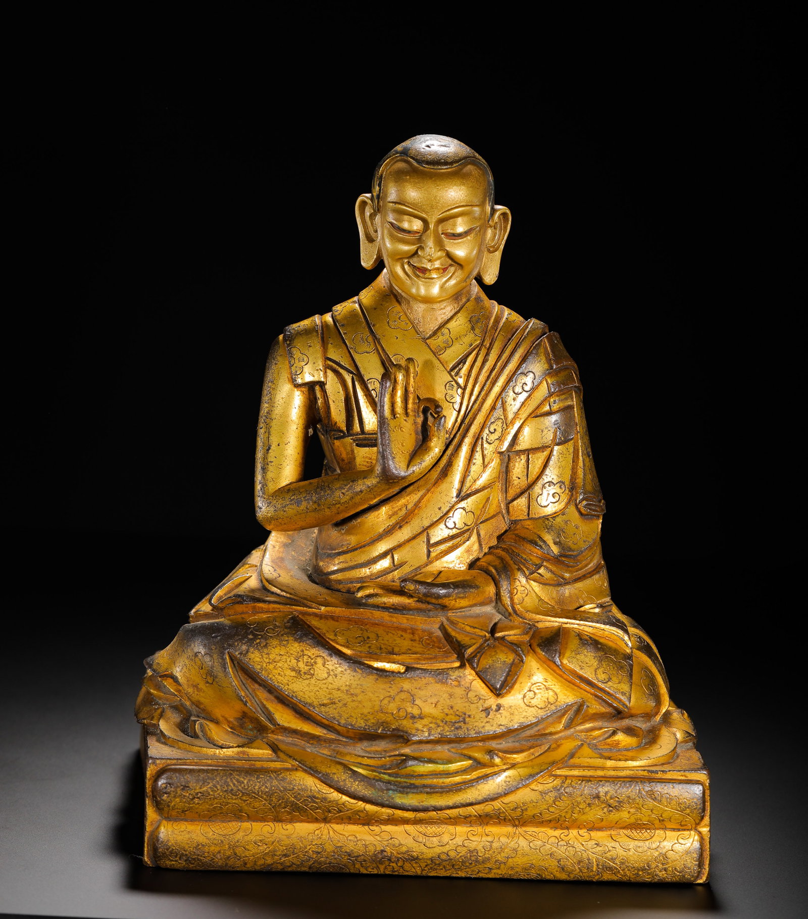 A GILDED BRONZE STATUE OF A MASTER FROM THE MING DYNASTY: H:24cm(9.45in)W:18cm(7.09in)Weight:5500g(12.13lb)Cast using the lost-wax method, the statue depicts the lama seated in lotus position on a base, with a warm, smiling face. The monastic robes are engra