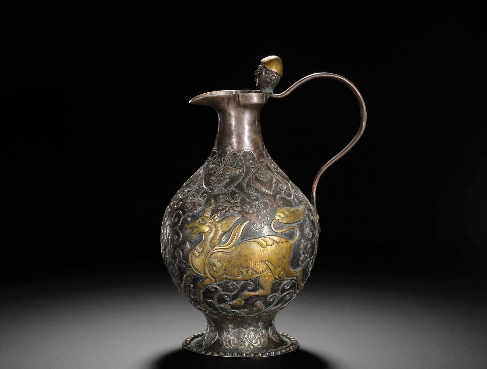 TANG DYNASTY SILVER-GILT EWER: HEIGHT:23CM (9.06IN) WIDTH:17CM (6.69IN) WEIGHT:610G (1.34LB) SILVER EWER WITH PARTIAL GILDING, TANG DYNASTY. ITS BODY IS FULLY CHASED WITH INTERLOCKING SCROLLS AND EMBOSSED WITH A GILDED FLORAL WINGE