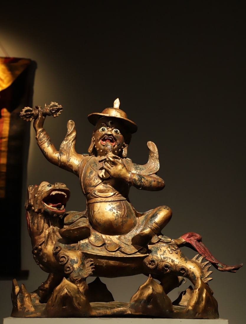 A LION-RIDING GUARDIAN STATUE FROM THE QING DYNASTY: WEIGHT:11150G.L:50CMH:49CMW:30CM THIS IS A TIBETAN BUDDHIST GILT-BRONZE STATUE OF THE DHARMA PROTECTOR DAMCHEN RIDING A LION. MADE OF GILT BRONZE, THE PROTECTOR HAS A FIERCE FACE WITH THREE EYES, RIDI