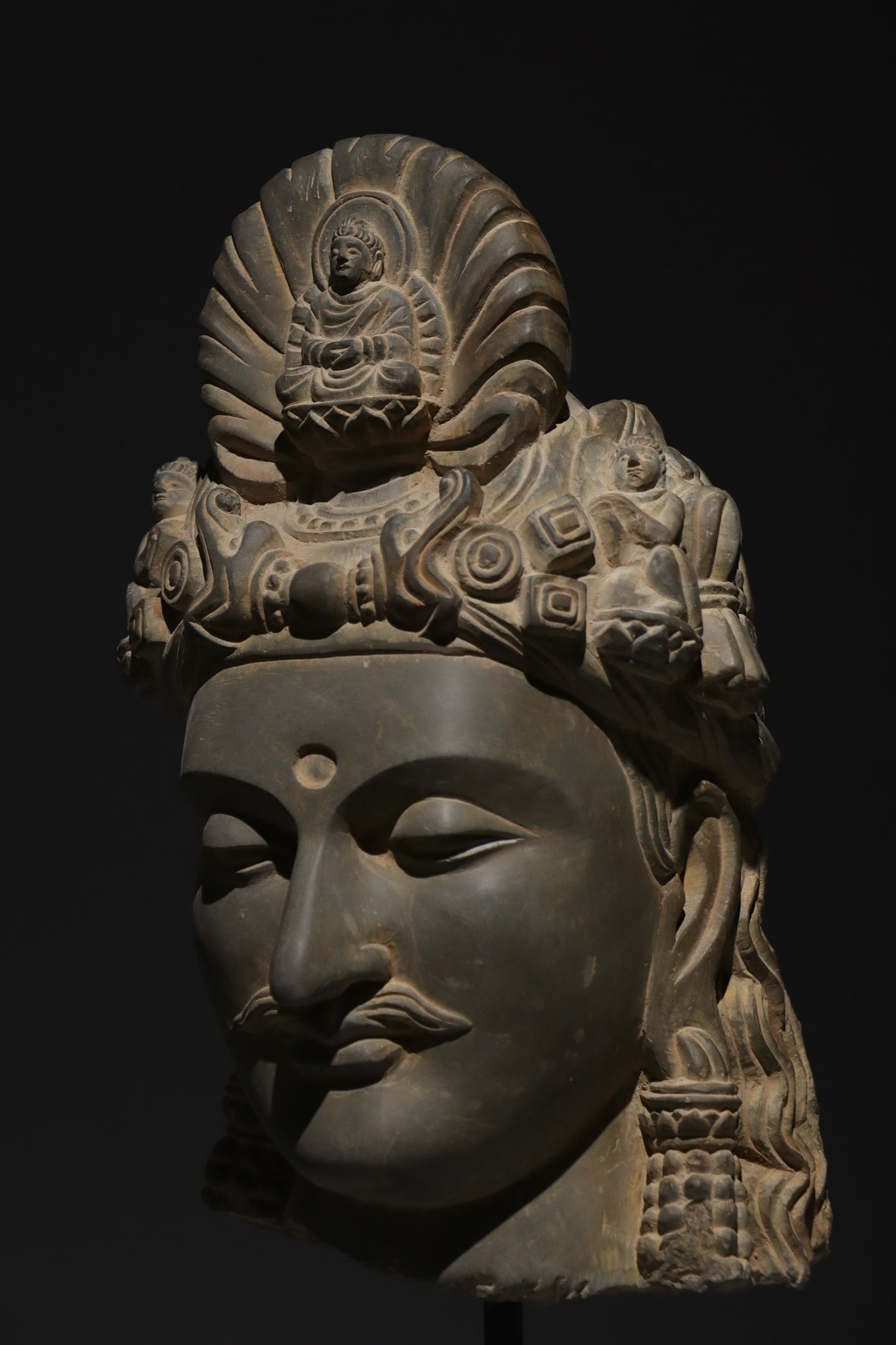 3RD CENTURY GANDHARA: H:56cm H:33cm This is a stone head of Guanyin Bodhisattva in Gandhara style, with a seated Buddha (a nimitta-buddha) carved at the center of its ornate crown. It features a serene and realistic face, 