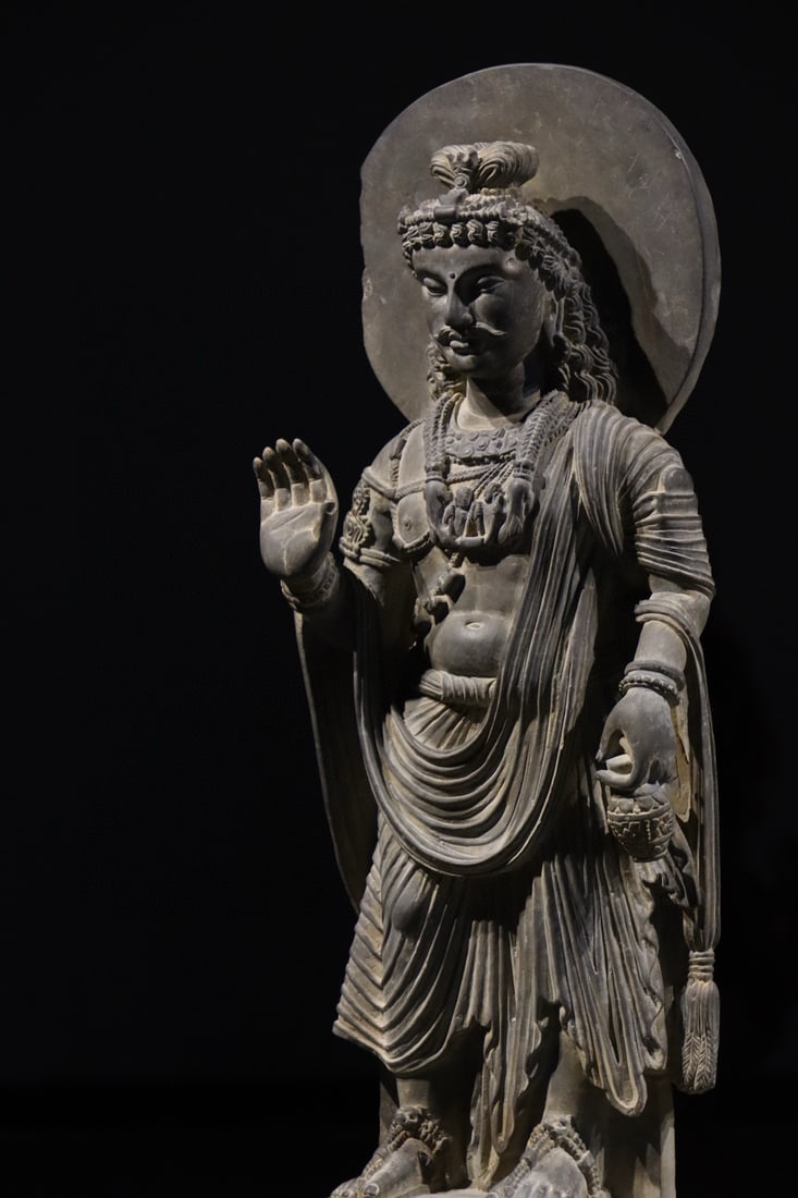 3RD CENTURY GANDHARA STATUES: H:61cm H:35cm W:45cm This is a standing Gandhara-style stone statue of Maitreya Bodhisattva, with the right hand forming Abhaya Mudra and the left hand holding a water bottle. Adorned with an ornate c