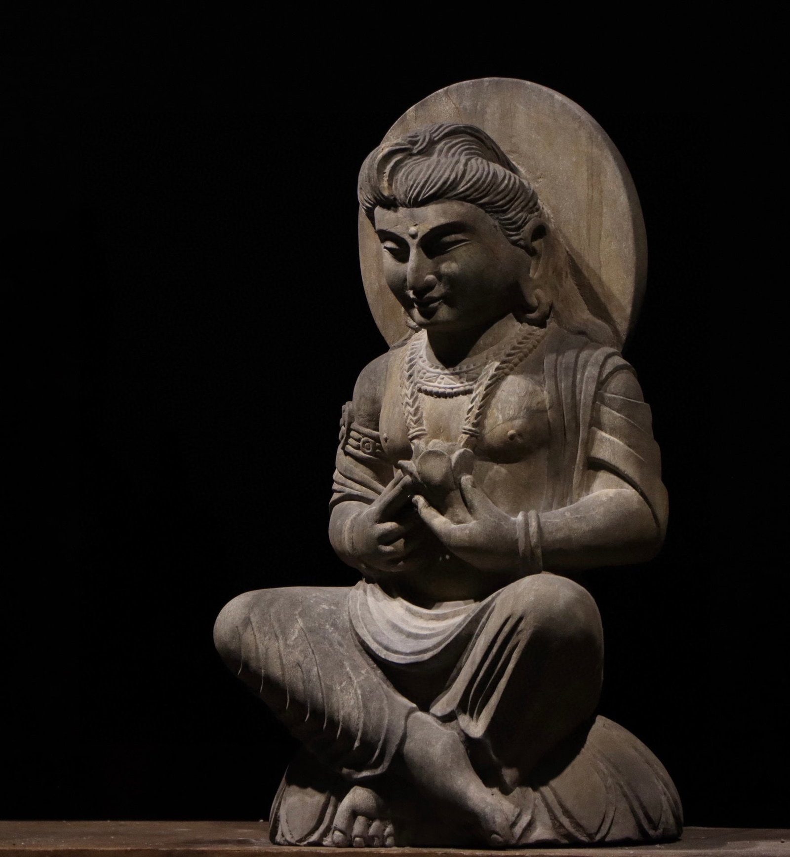 3RD CENTURY ANCIENT GREEK GANDHARA STATUES: H:69cm W:37cm T:33cm This is a seated stone statue of a Bodhisattva in Gandhara style, holding a lotus flower with both hands and smiling. It features a circular halo behind, representing a classic re