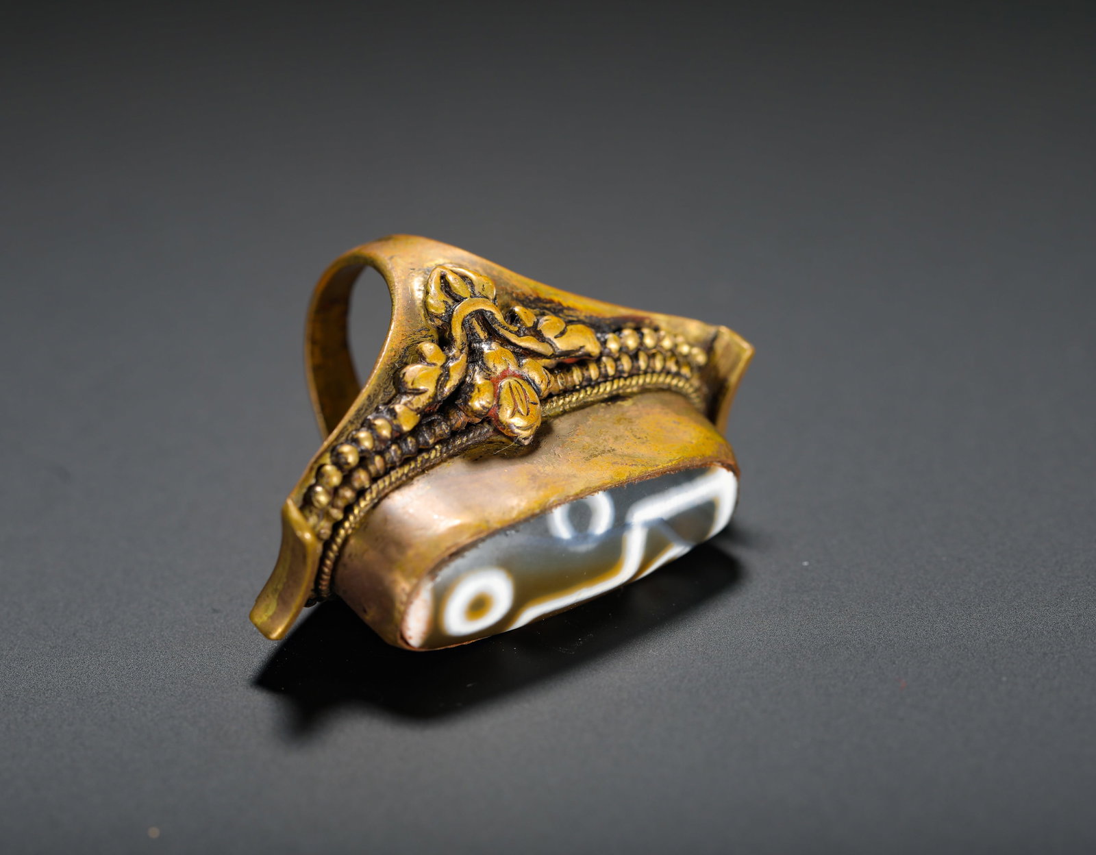 COPPER GILT EMBOSSED FLORAL SET WITH SIX-EYED SKY BEADS RING: Height:4.6cm (1.81in) D:3.5cm (1.38in) Weight:28g(0.06lb)Gilt bronze ring embossed with scrolling flowers inlaid with six-eye dzi bead. The bezel is carved with interlocking floral and bead patterns, 
