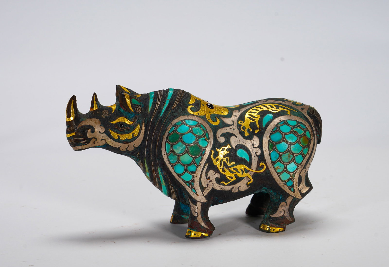 BRONZE FROM THE WARRING STATES PERIOD WITH INLAID GOLD AND SILVER AND TURQUOISE RHINOCEROS: Height:8cm (3.15in) Width:14.5cm (5.71in) Weight:500g(1.10lb)Solid bronze zoomorphic sculpture of rhinoceros. Decorated with interlaced golden tiger-dragon patterns, turquoise scale inlay, mixed gold 