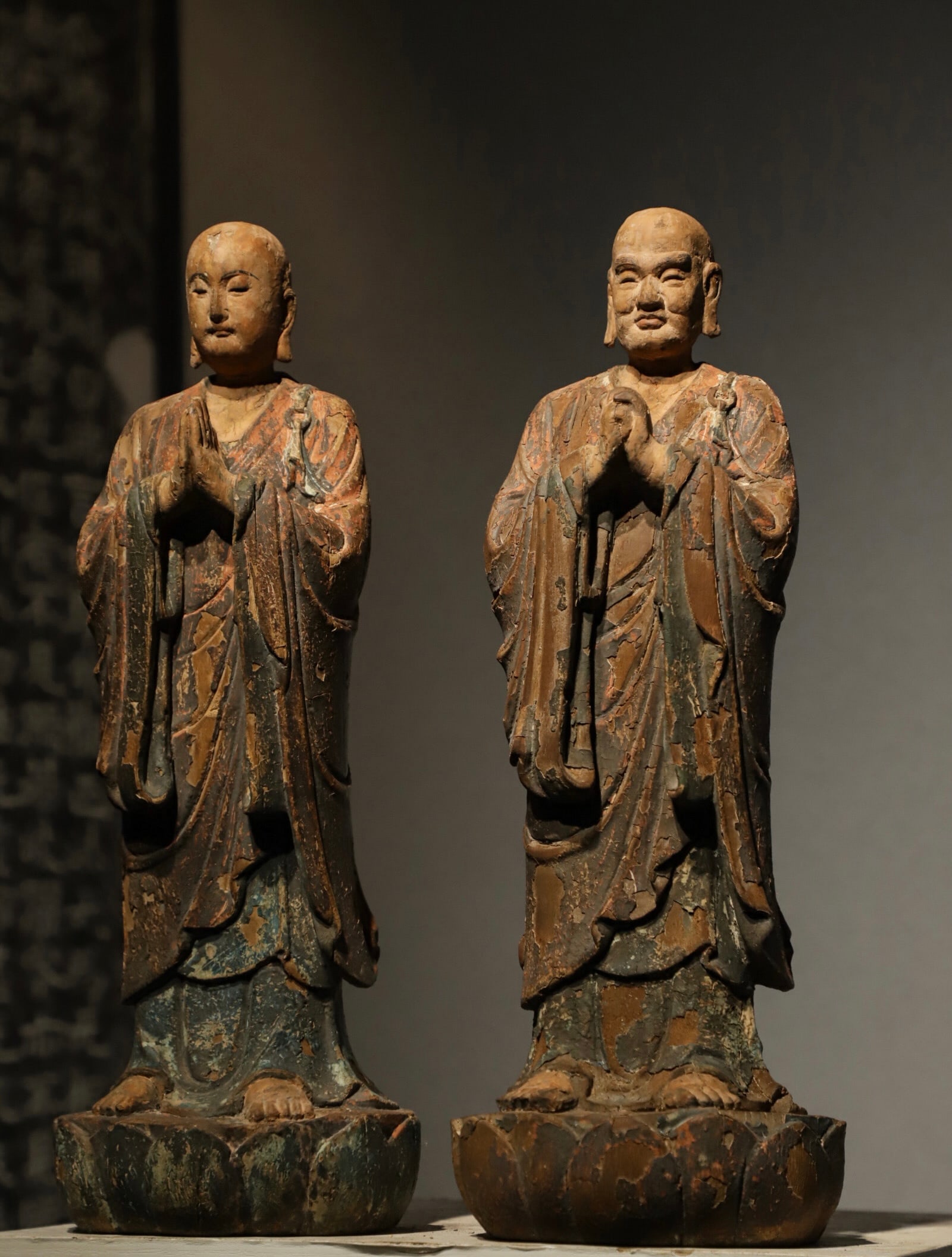 A PAIR OF STANDING STATUES OF VENERABLE ANANDA KASYAPA IN PAINTED WOODCARVING FROM THE MING DYNASTY: H:46cm (18.11in):Two solid wood-carved attendant arhat statues painted with mineral pigments; aged lacquer in red, blue and brown peels partially off. Standing separately on lotus plinths as Shakyamun