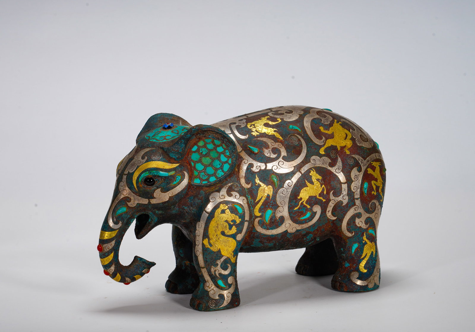 BRONZE STATUE WITH INLAID GOLD AND SILVER AND TURQUOISE FROM THE WARRING STATES PERIOD: Height:15cm (5.91in) Width:25cm (9.84in) Weight:2980g(6.57lb)Solid bronze sculpture of standing elephant. Body decorated with interlaced tiger-phoenix scrolls in gold and silver wire, turquoise mosaic