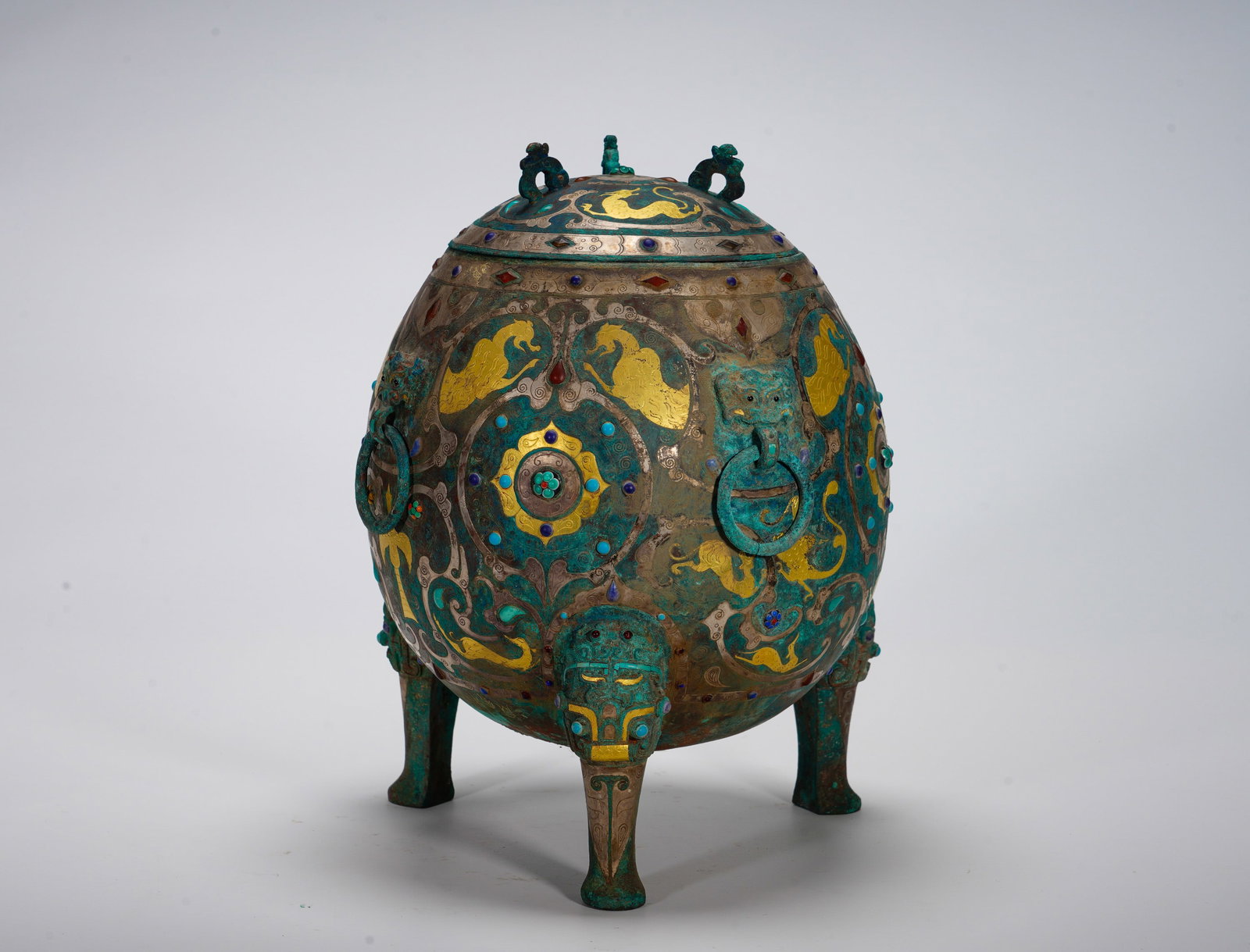 A FOOTBALL-SHAPED COVERED TRIPOD INLAID WITH GOLD AND SILVER INLAID WITH MYTHICAL BEASTS FROM THE: Height:7cm (2.76in) Width:21cm (8.27in) Weight:3140g(6.92lb)Globular covered sacrificial vessel supported by three beast-head feet, fitted with beast-ring handles. Surface decorated with galloping hor