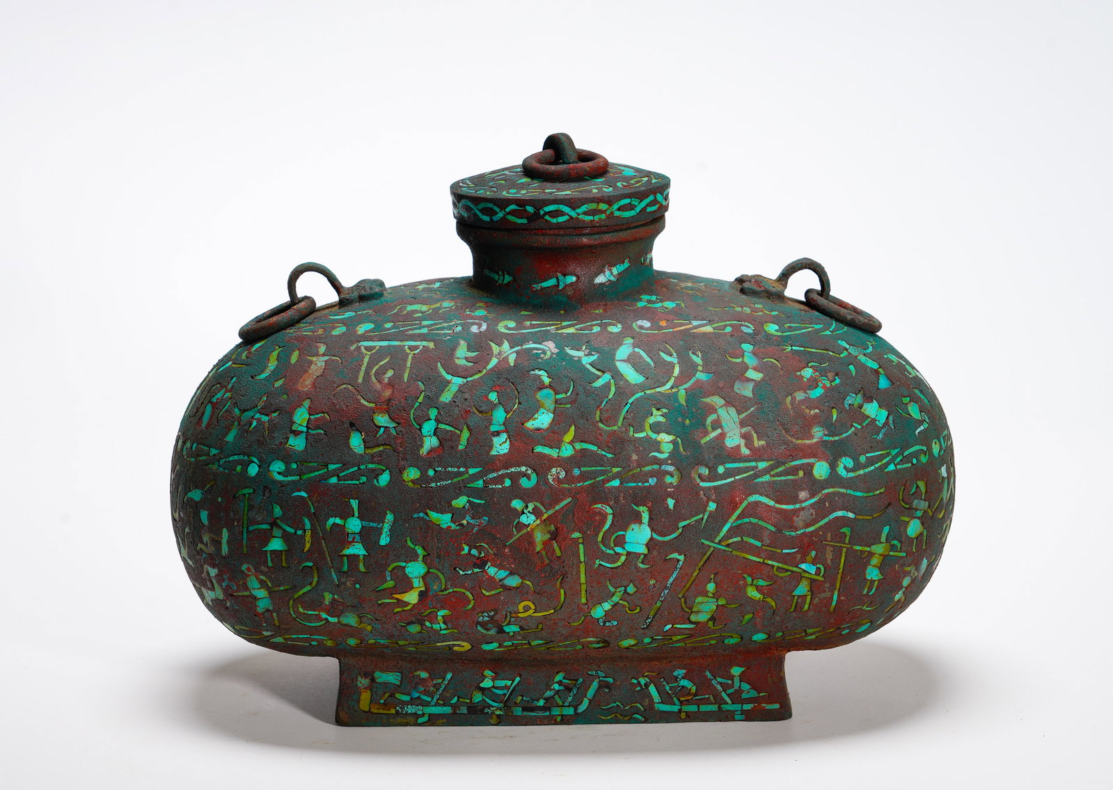 A FLAT POT WITH TURQUOISE INLAID PATTERNS OF WATER AND LAND BATTLES FROM THE WARRING STATES PERIOD: Height:22cm (8.66in) Width:30cm (11.81in) Weight:3220g(7.10lb)Flattened bronze covered wine vessel with ring handles. Continuous horizontal bands inlaid with turquoise depict warriors, combat, hunt an