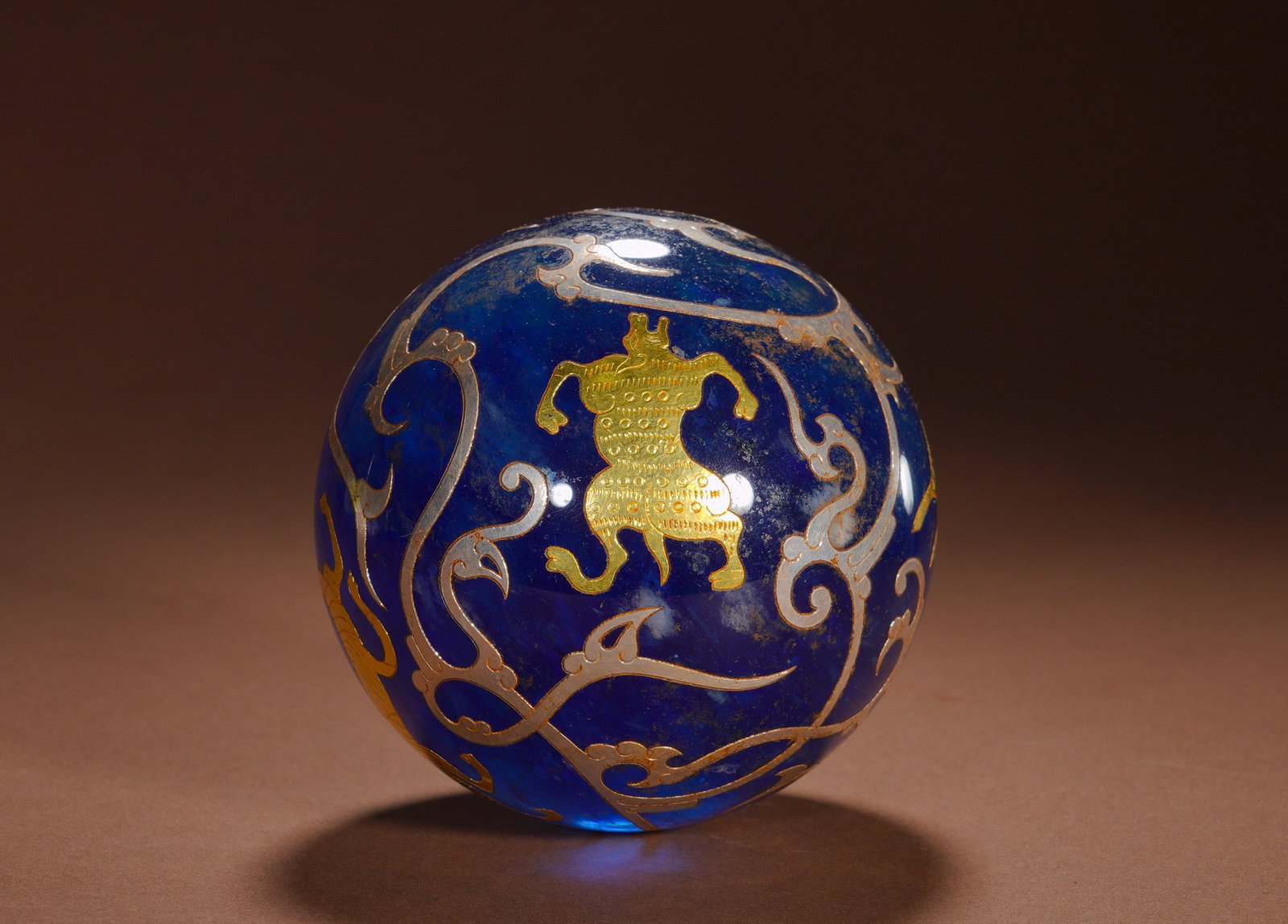 A ROUND BALL WITH INLAID GOLD AND SILVER PATTERNS OF GODS AND AUSPICIOUS BEASTS FROM THE HAN DYNASTY: Diameter:9.5cm (3.74in) Weight:1100g(2.42lb)Cast ancient glass sphere in deep sapphire tone. Gilded relief immortal figure decorates the surface, intertwined phoenix and scroll patterns outlined by si