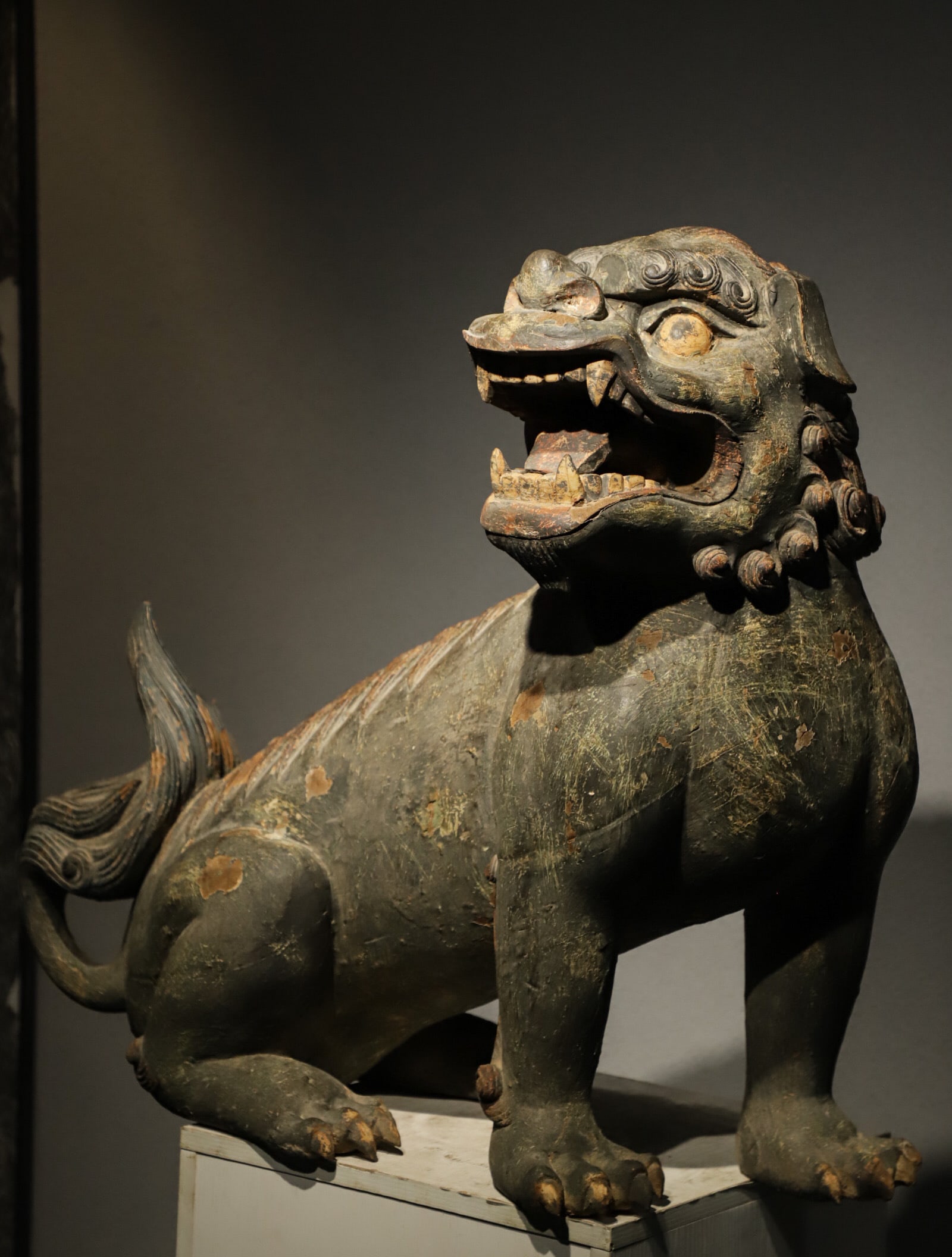 A PAINTED CROUCHING LION FIGURINE CARVED FROM WOOD FROM THE MING DYNASTY: L:80cm (31.50in) W:40cm (15.75in) H:70cm (27.56in)Solid wood carved auspicious lion coated with mineral green and ochre pigments; finely detailed fur patterns, partially peeling lacquer revealing wood