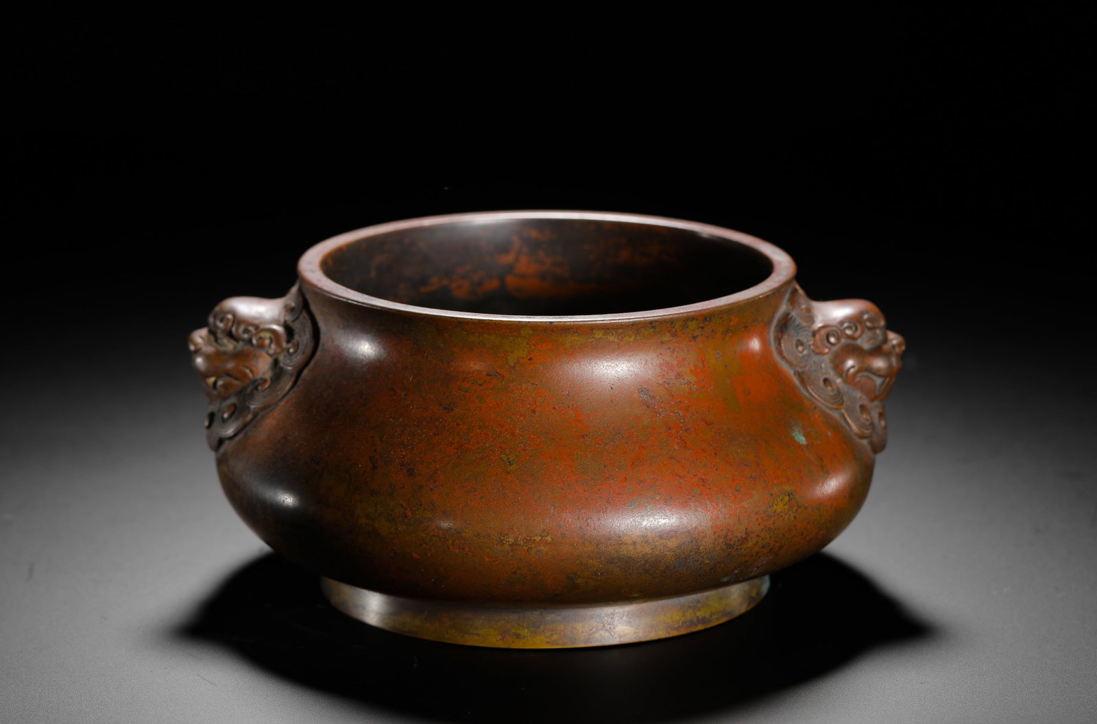 BRONZE LION-EAR STOVE OF THE QING DYNASTY: H:7cm (2.76in) W:16cm (6.30in) Weight:1220g (2.69lb) This is a Qing Dynasty bronze incense burner with a flattened body and lion-head handles on both sides. The base is inscribed with the seal script 