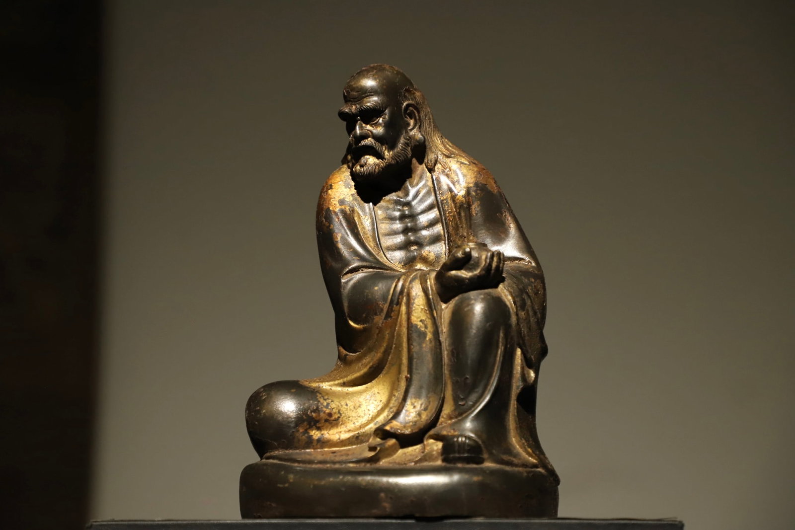 A GILDED BRONZE STATUE OF BODHIDHARMA FROM THE MING DYNASTY: Height:23cm(9.06in), Long:15cm(5.91in), Width:12cm(4.72in)This gilt bronze seated statue of Bodhidharma dates to the Ming Dynasty. Crafted in gilt bronze, its gilded surface bears mottled wear, exudin