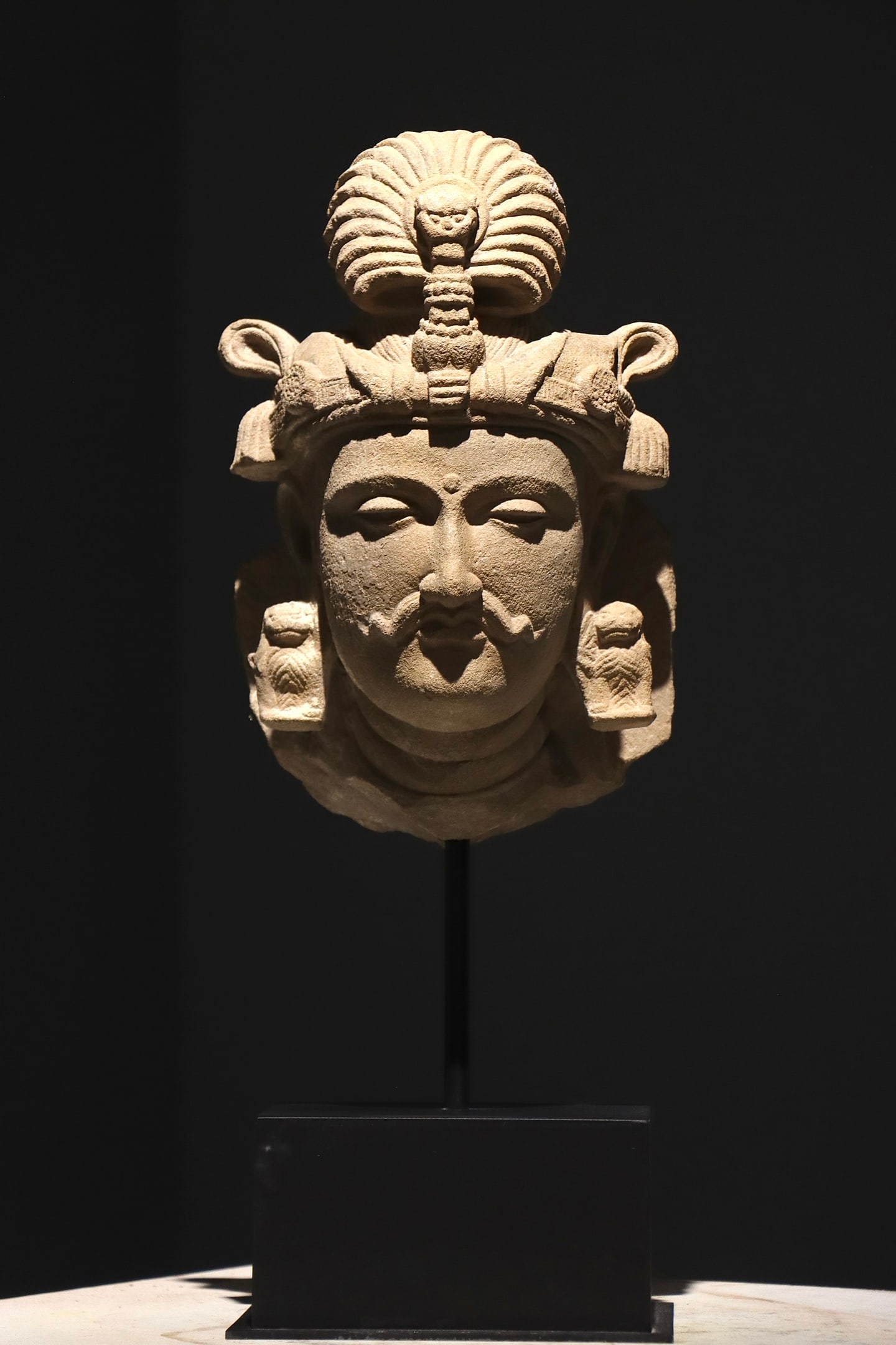 4TH CENTURY GANDHARA-STYLE SANDSTONE GUARDIAN: Height:54cm(21.26in), Height:33cm(12.99in)Sandstone deity head in classic Gandhara style. The sculpture wears an ornate crown decorated with serpent motif on top. It features an urna on the forehead, 