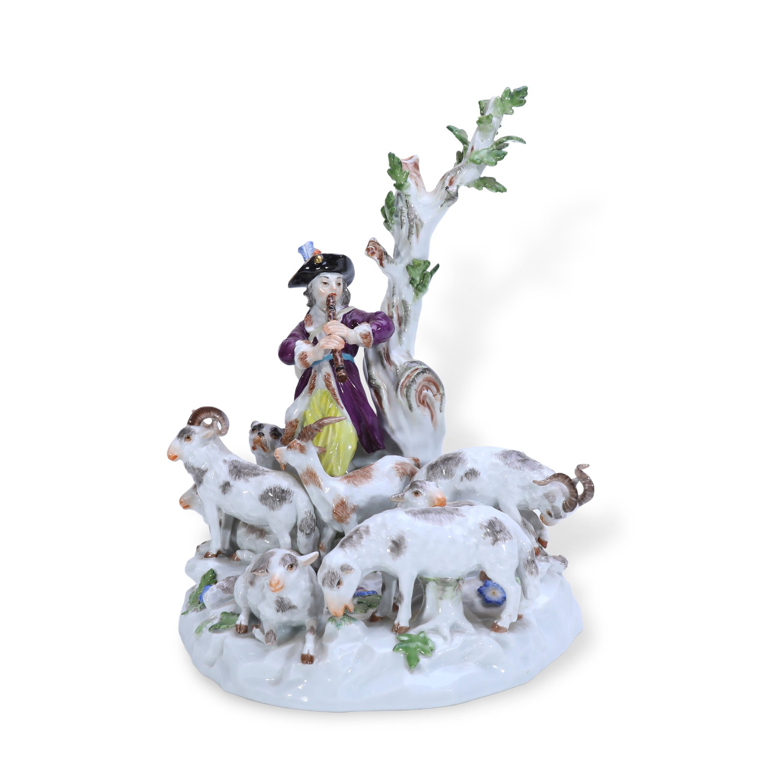 19th Century Meissen Porcelain Group Figurine of Piper Shepherd with Flock of Sheep: H:23.5cm W:18.5cm A young shepherd wearing a tricorn hat leans against a withered tree playing a flute. Multiple goats (rams, ewes and lambs) surround the base, with wild flowers and foliage scattered