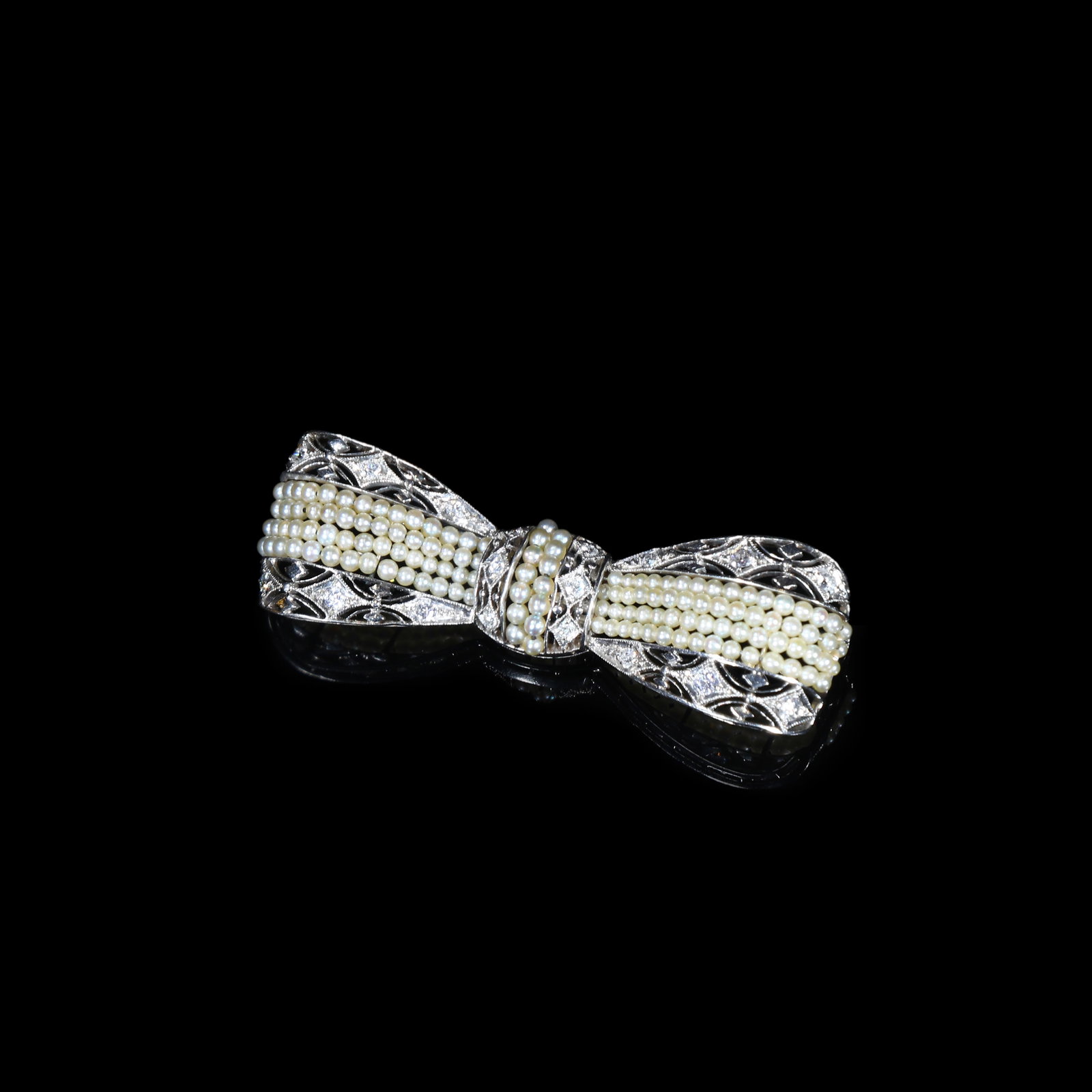 Edwardian-Period (Circa 1900-1915) White-Gold Natural-Pearl Bow Brooch: 1.7*5.4cm Wt:10.4g Edwardian-period (circa 1900-1915) bow brooch in white-gold. Designed as a bow with open-work filigree, multiple strands of natural pearls form the bow loops, the metal finely chase