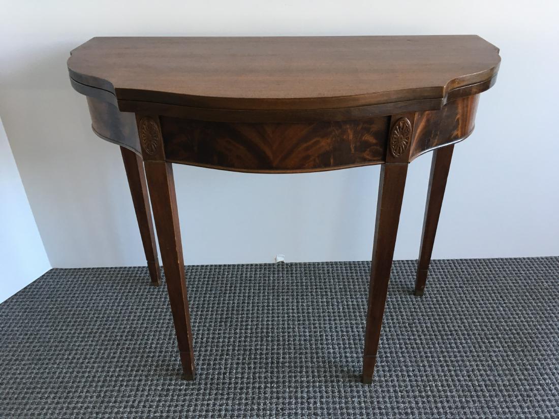 MAHOGANY HEPPLEWHITE STYLE GAME TABLE: Mahogany Hepplewhite style game table with serpentine front, swivel top, tapered legs, applied carved oval decoration on flamed mahogany veneer frieze - 35.75" l x 18" w x 29.75" h (left brass end cap