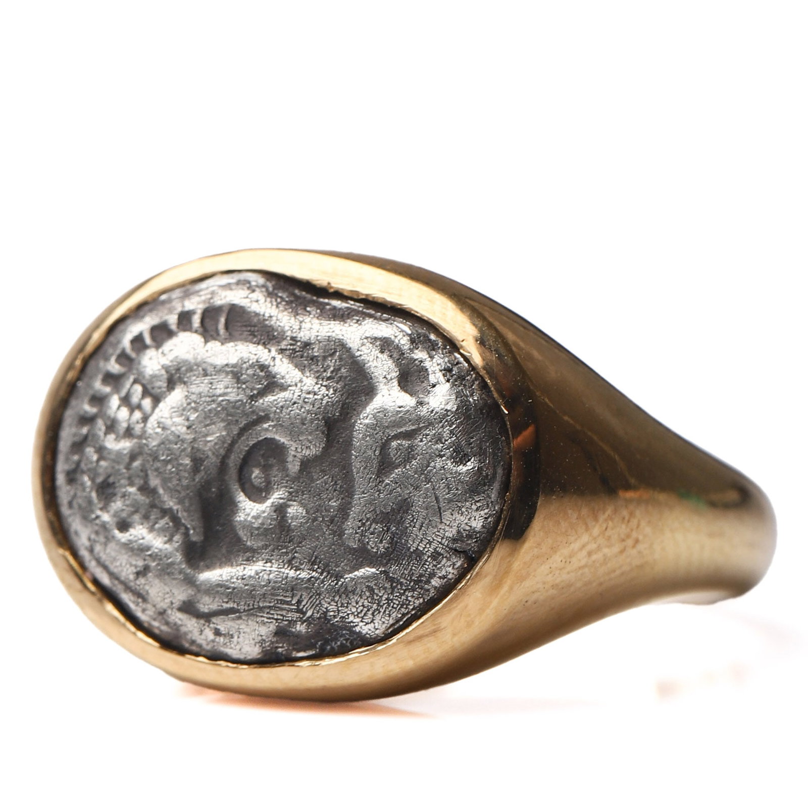 A GOLD RING SET WITH AN ANCIENT SILVER COIN: Dimensions Inner diameter 17 mm; face 9/16 × 7/16 in (14.6 × 11 mm); weight approx. 13.1 g. Description: The oval bezel mounted with an ancient silver coin, possibly Greek or Hellenistic, depicting 