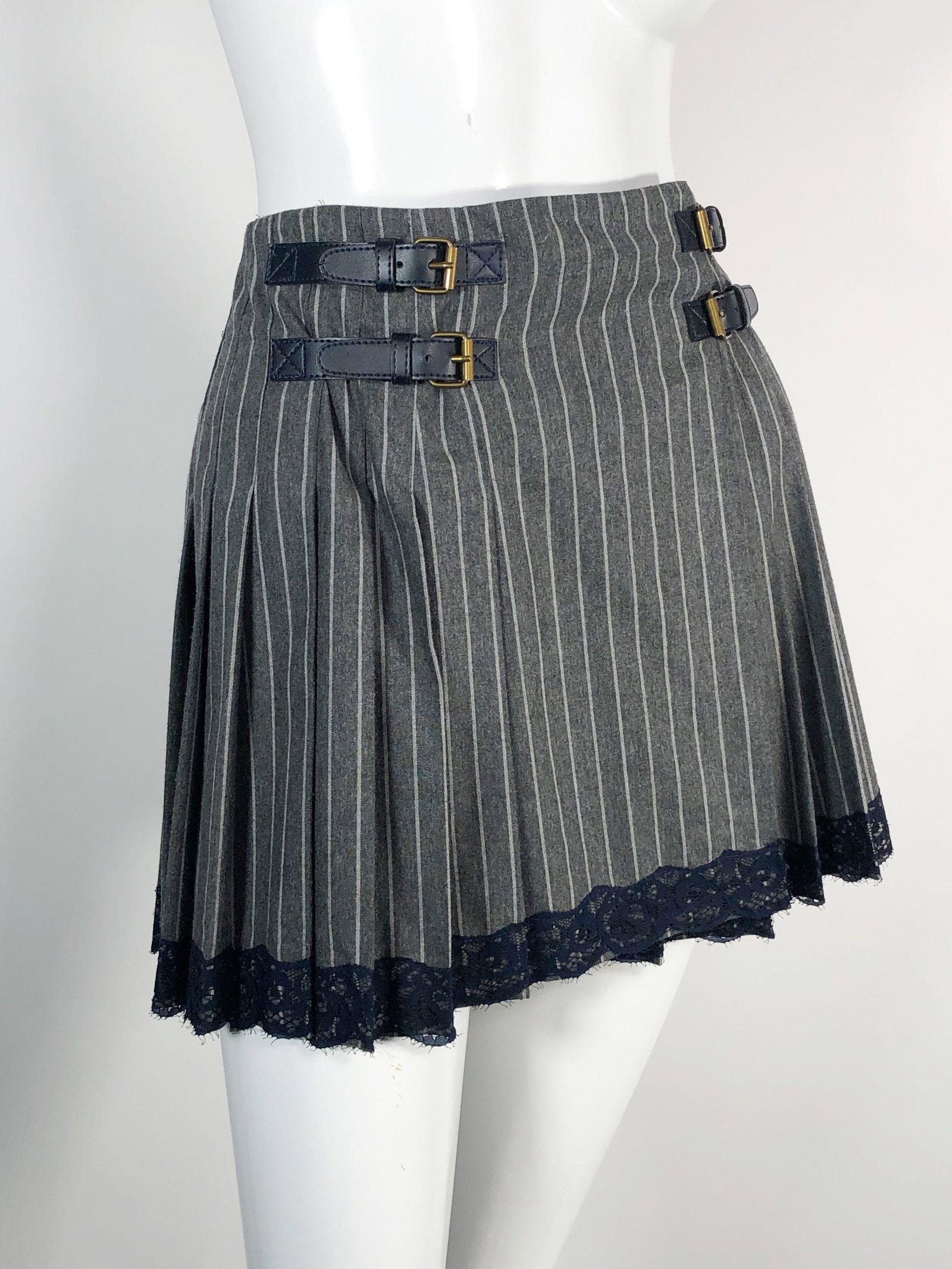 John Galliano Grey Pinstripe Pleated Kilt Mini Skirt: John Galliano grey pinstripe pleated rayon/wool mini skirt with kilt-style buckles at hips and black lace trim at hem. Size 38 Italian, Size US small.