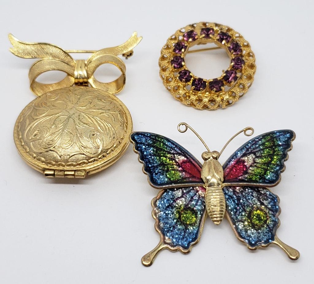 3-GOLD TONED BROOCHES: (1)AVON PERFUME LOCKET: (1)GOLD TONED CIRCULAR BROOCH WITH VIOLET RHINESTONES & (1)BUTTERFLY BROOCH WITH ENAMEL