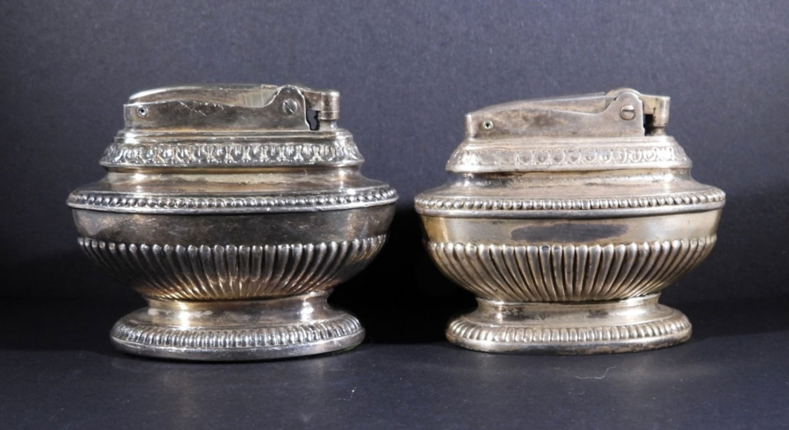 (2) RONSON QUEEN ANNE TABLE LIGHTERS: TWO RONSON TABLE LIGHTERS. ONE IS CLEARLY MARKED "QUEEN ANNE". THE OTHER IS NOT MARKED WITH A NAME AND IS SLIGHTLY DIFFERENT. SILVER TONE, POSSIBLY SILVER PLATED. 3.5" WIDE, 2.5-3" TALL.